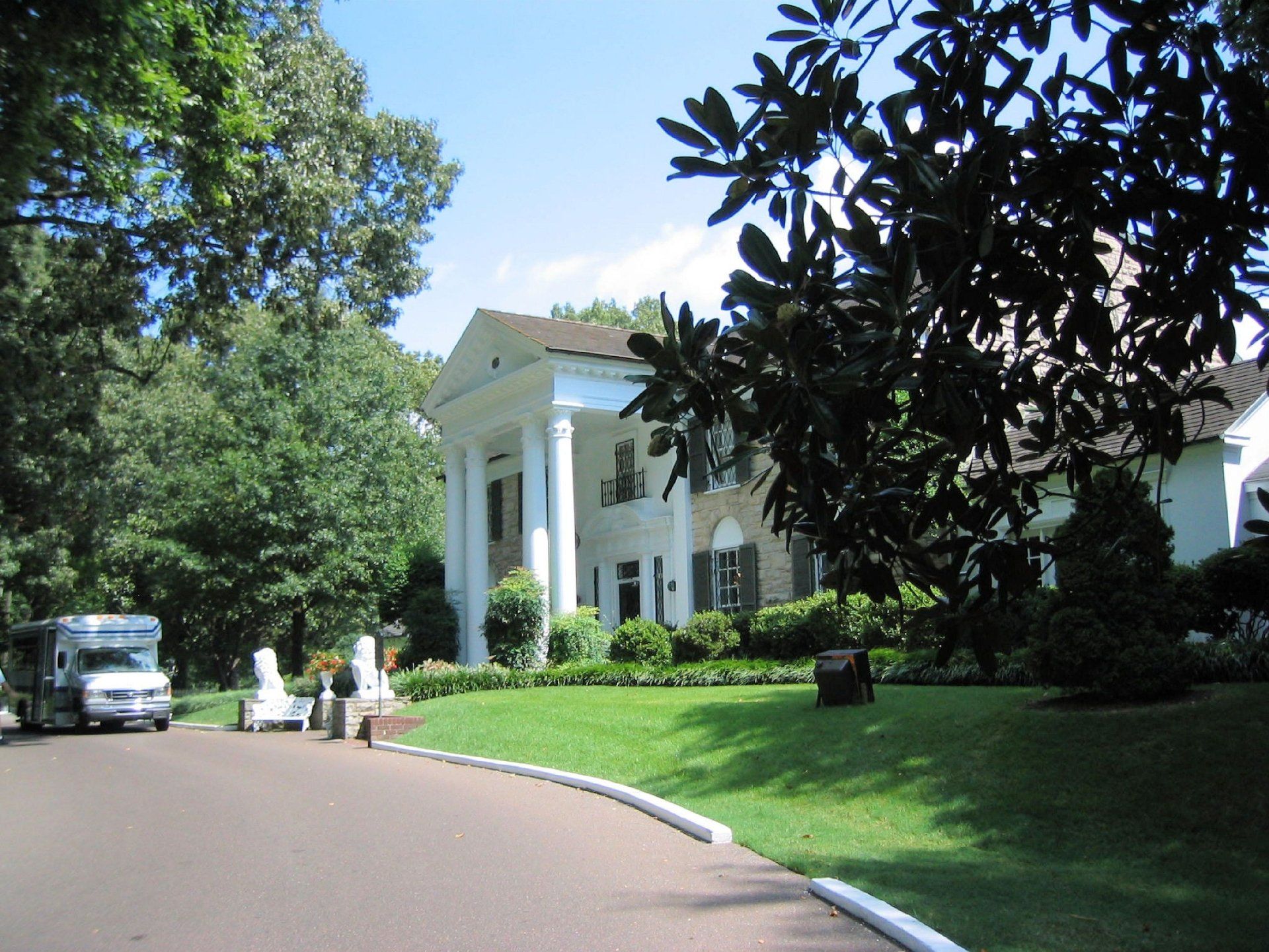 Landhuis Graceland met witte zuilen, een groen gazon, een blauwe lucht en een busje.