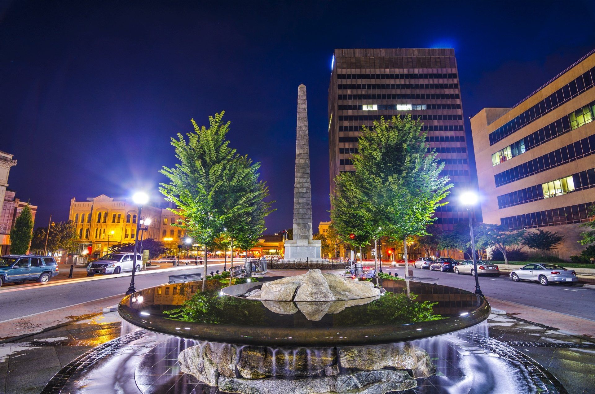 Nachtzicht op een stadsplein met een fontein, obelisk, gebouwen, straatverlichting en auto's.