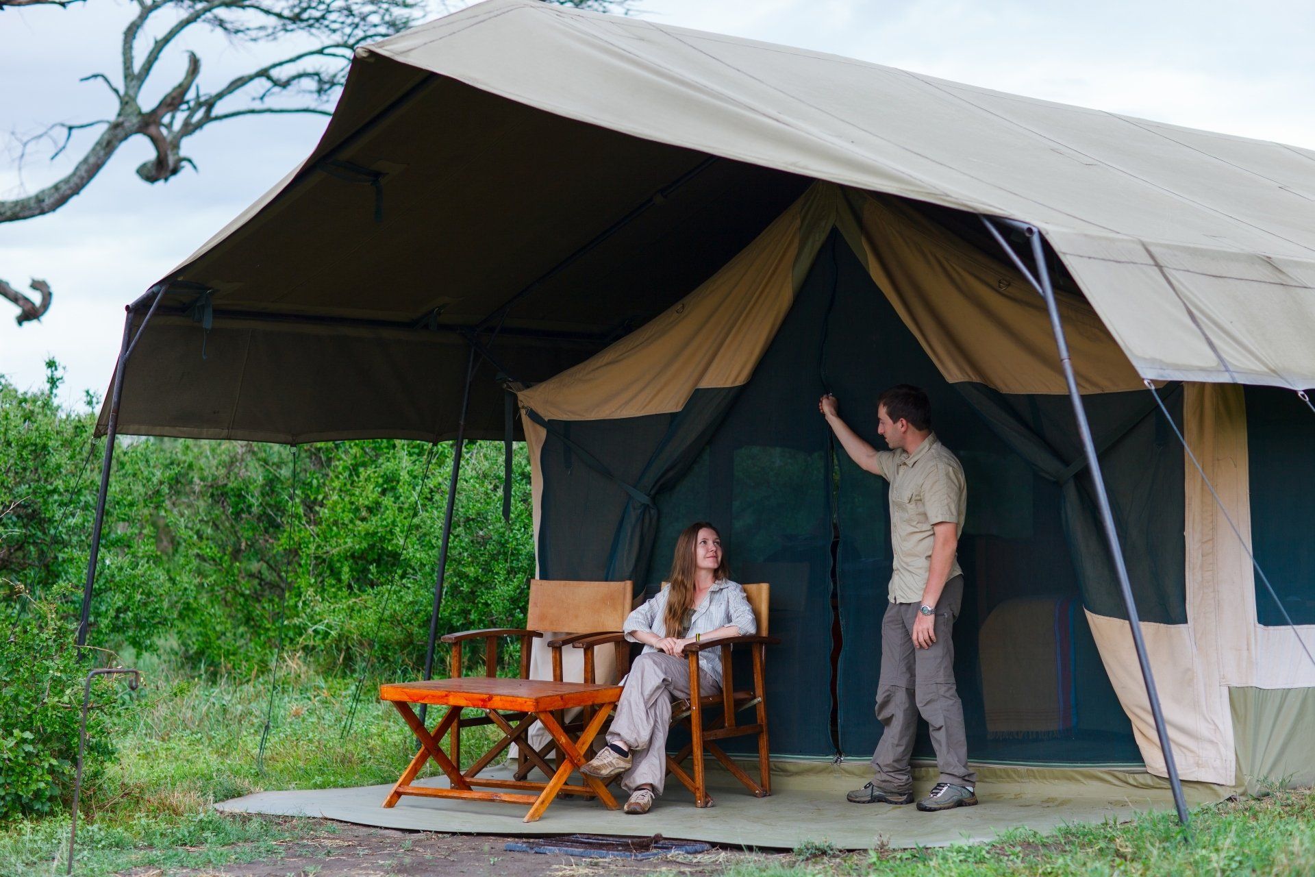 Een stel staat bij de ingang van de tent, de een zit, de ander staat, en kijkt naar binnen. Een kaki-bruine tent in een grasrijke omgeving.