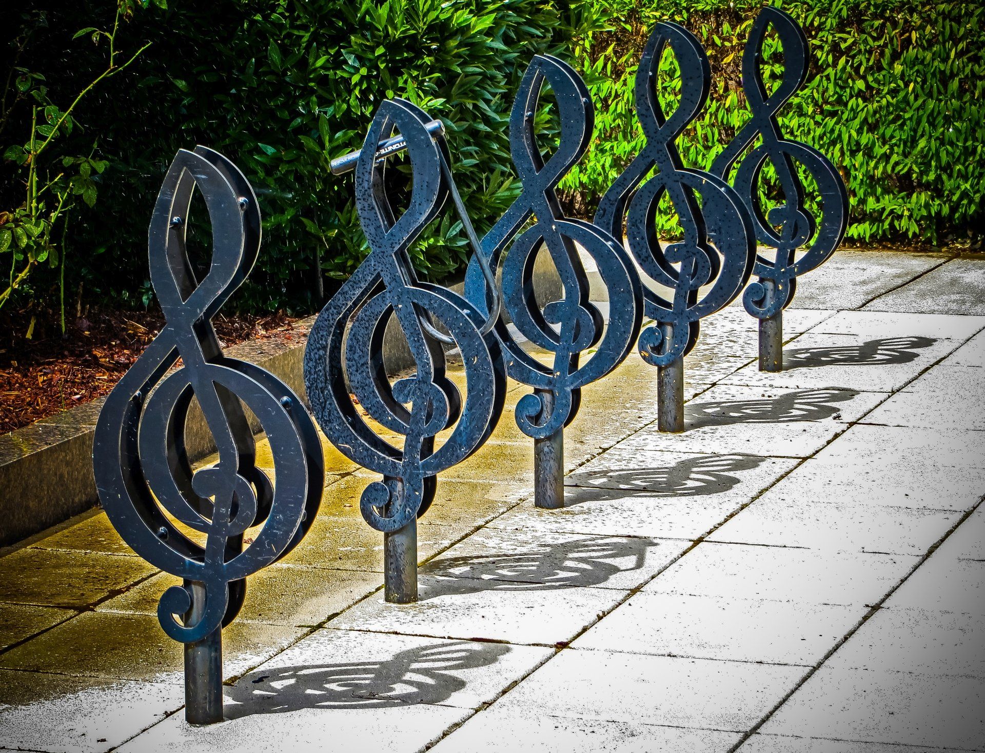 Vijf zwarte metalen vioolsleutelsculpturen op een geplaveid pad, die schaduwen werpen.
