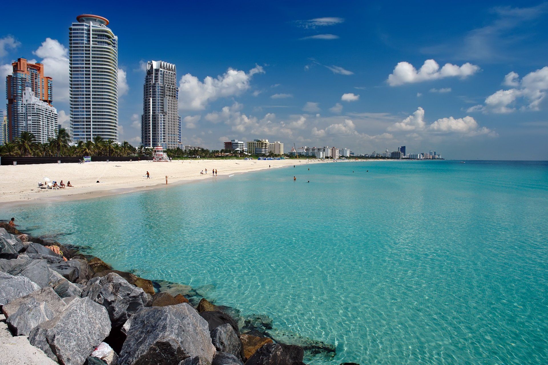 Een strand met turquoise water, wit zand en hoge gebouwen onder een blauwe hemel met wolken.
