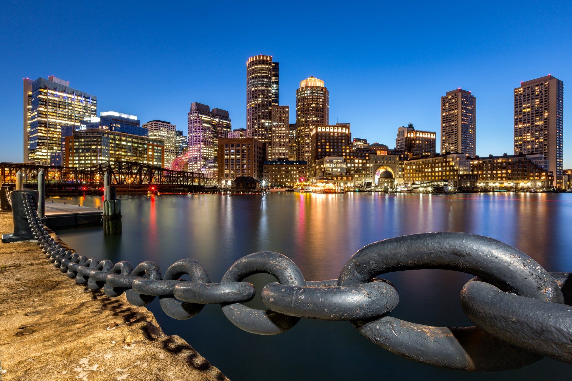 Nachtelijk uitzicht op de skyline van Boston met verlichte gebouwen, water op de voorgrond en een grote ketting op de voorgrond.