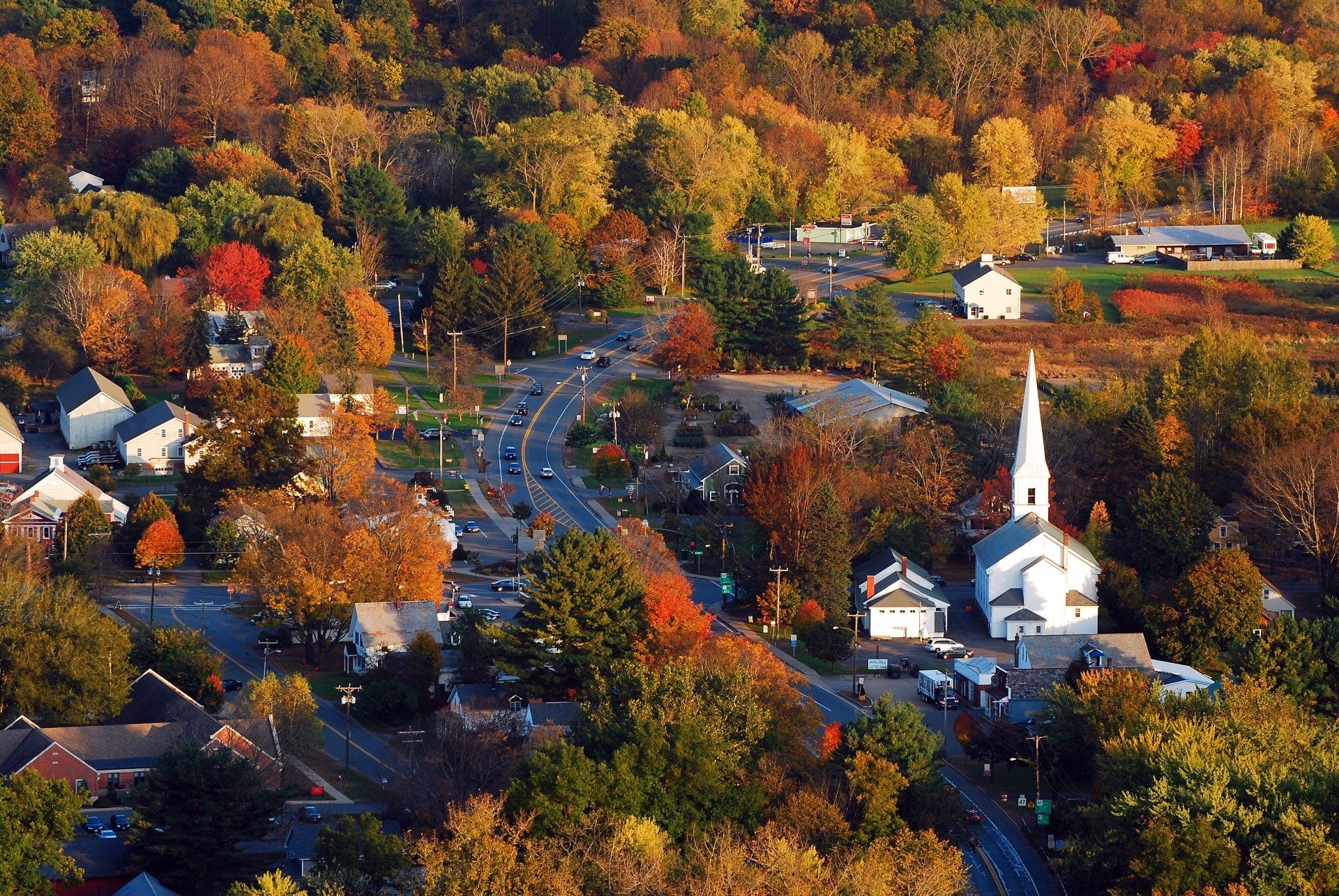Luchtfoto van een stad in de herfst; een kronkelende weg met auto's, omgeven door bomen met kleurrijk gebladerte, een witte kerk met torenspits.