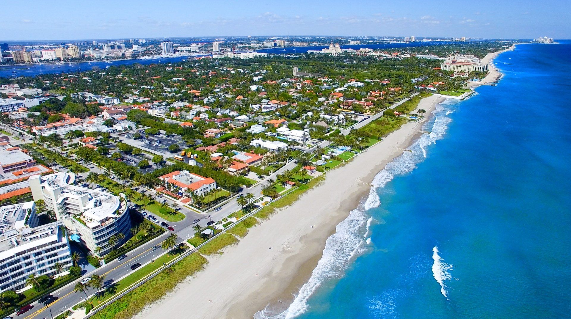 Luchtfoto van een kuststad met strand, oceaan, gebouwen en weelderige groene bomen onder een helderblauwe hemel.