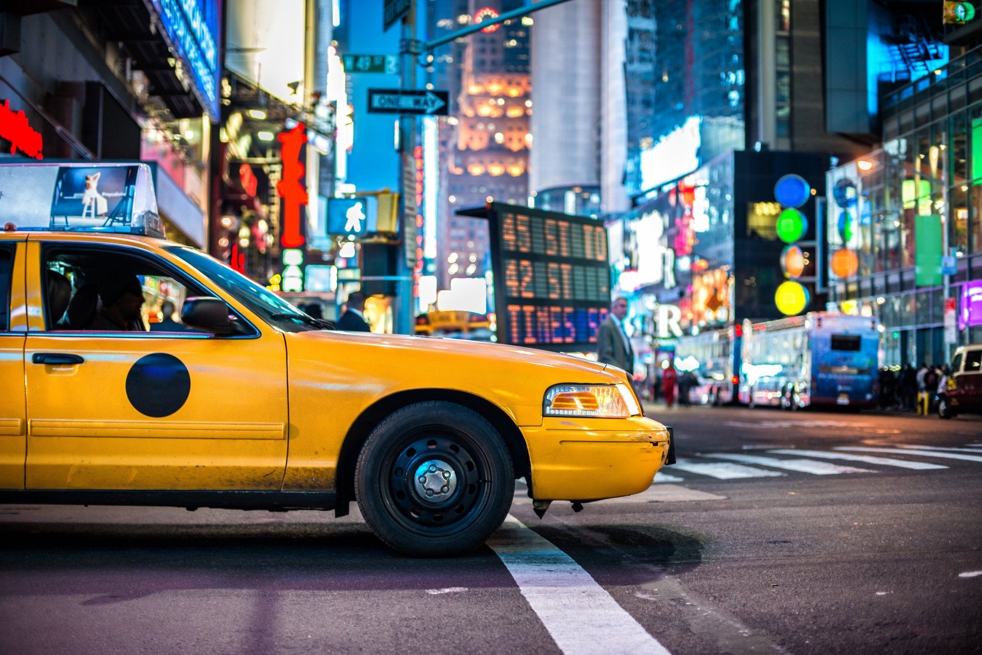 Een gele taxi in een stadsstraat, met felle koplampen en een blauw verlicht stadsbeeld.