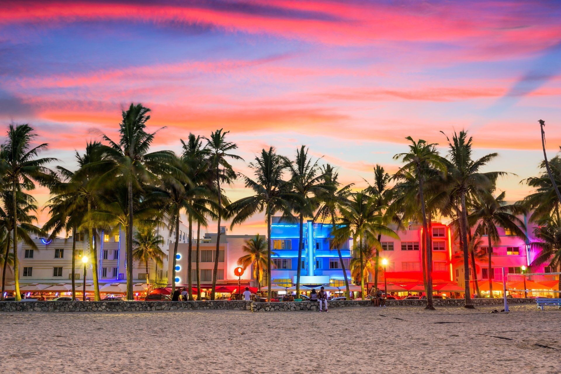 Uitzicht vanaf het strand op Miami bij zonsondergang; silhouetten van palmbomen tegen een kleurrijke hemel en verlichte art-decogebouwen.