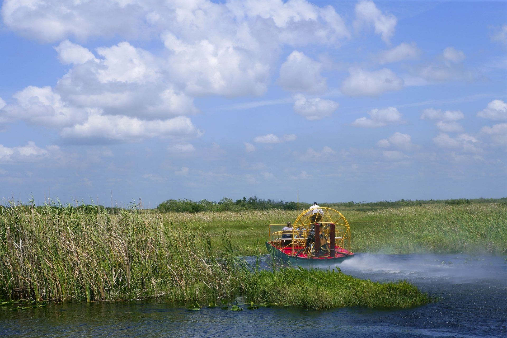 Een luchtboot scheurt over het moeras van de Everglades onder een blauwe hemel met wolken.