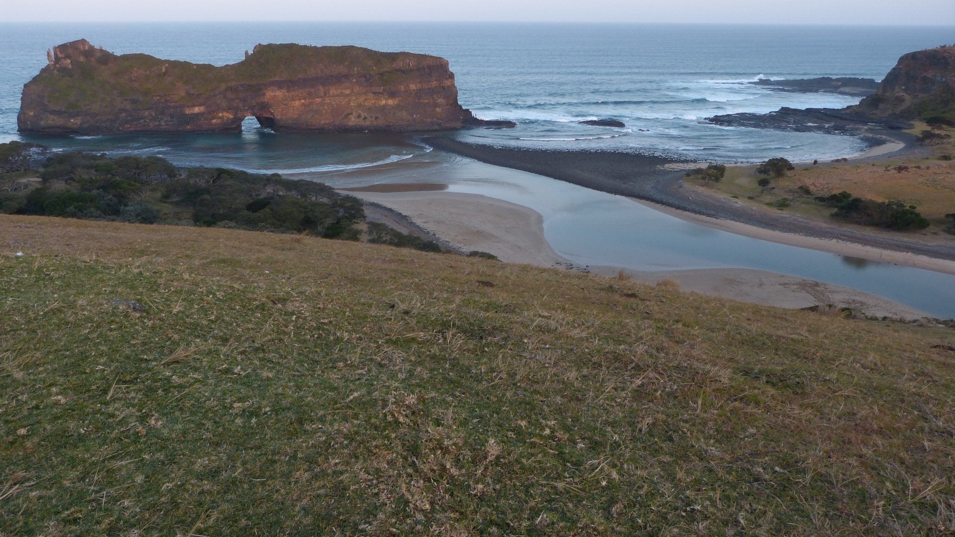 Een kustlandschap met een rotsachtige uitloper, een zandstrand en een rivier die onder een bleke hemel in de oceaan uitmondt.