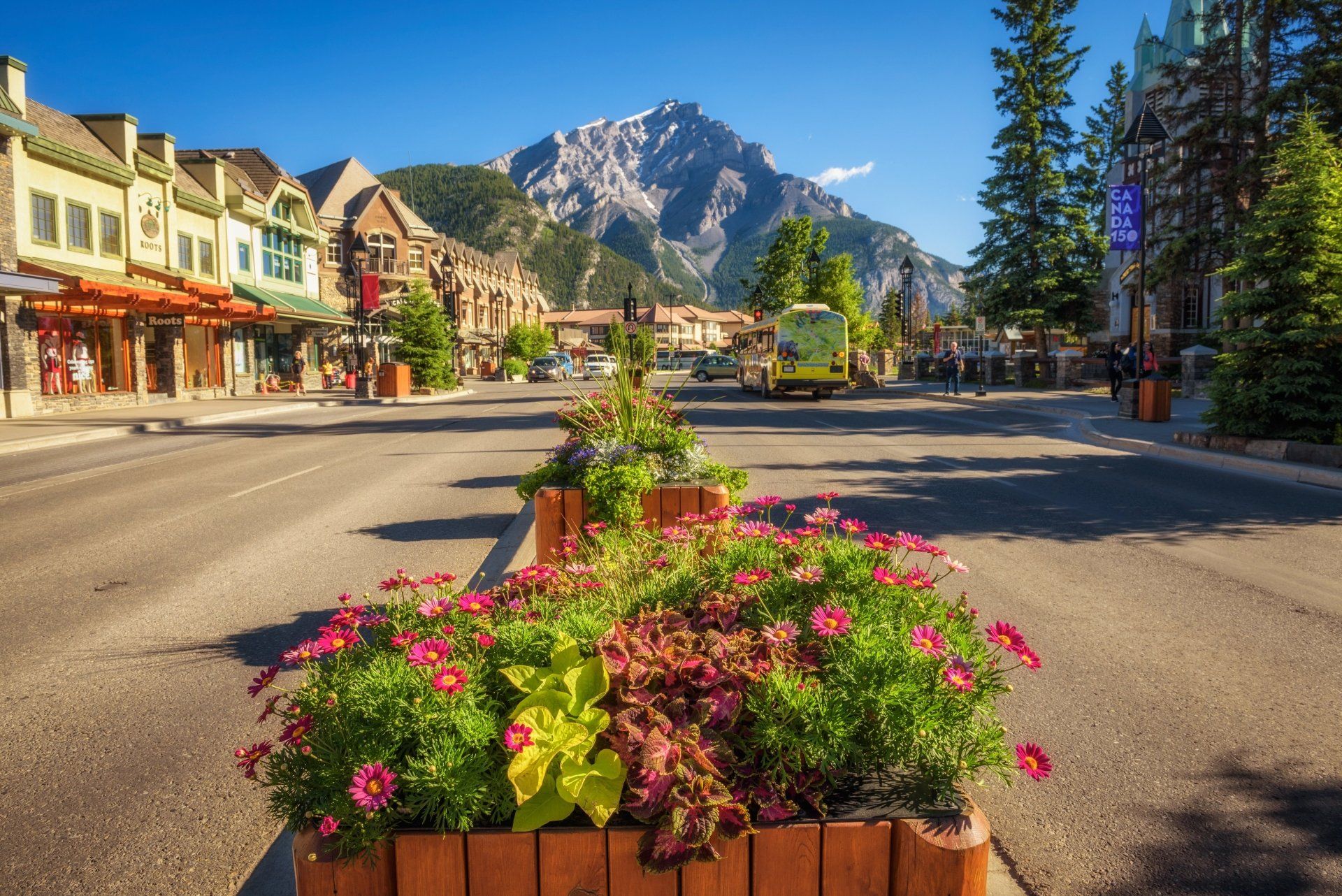 Straatbeeld in Banff, Canada, met winkels, bloembakken op de voorgrond en bergen in de verte onder een blauwe hemel.