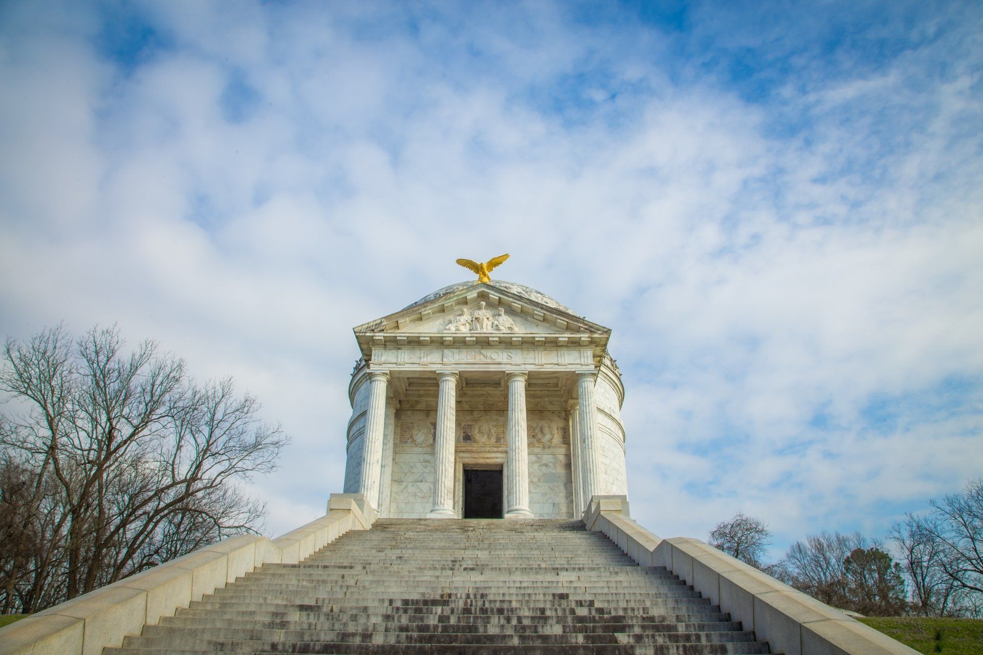 Wit neoklassiek mausoleum bovenop stenen trappen, gouden adelaar op het dak onder een bewolkte blauwe hemel.