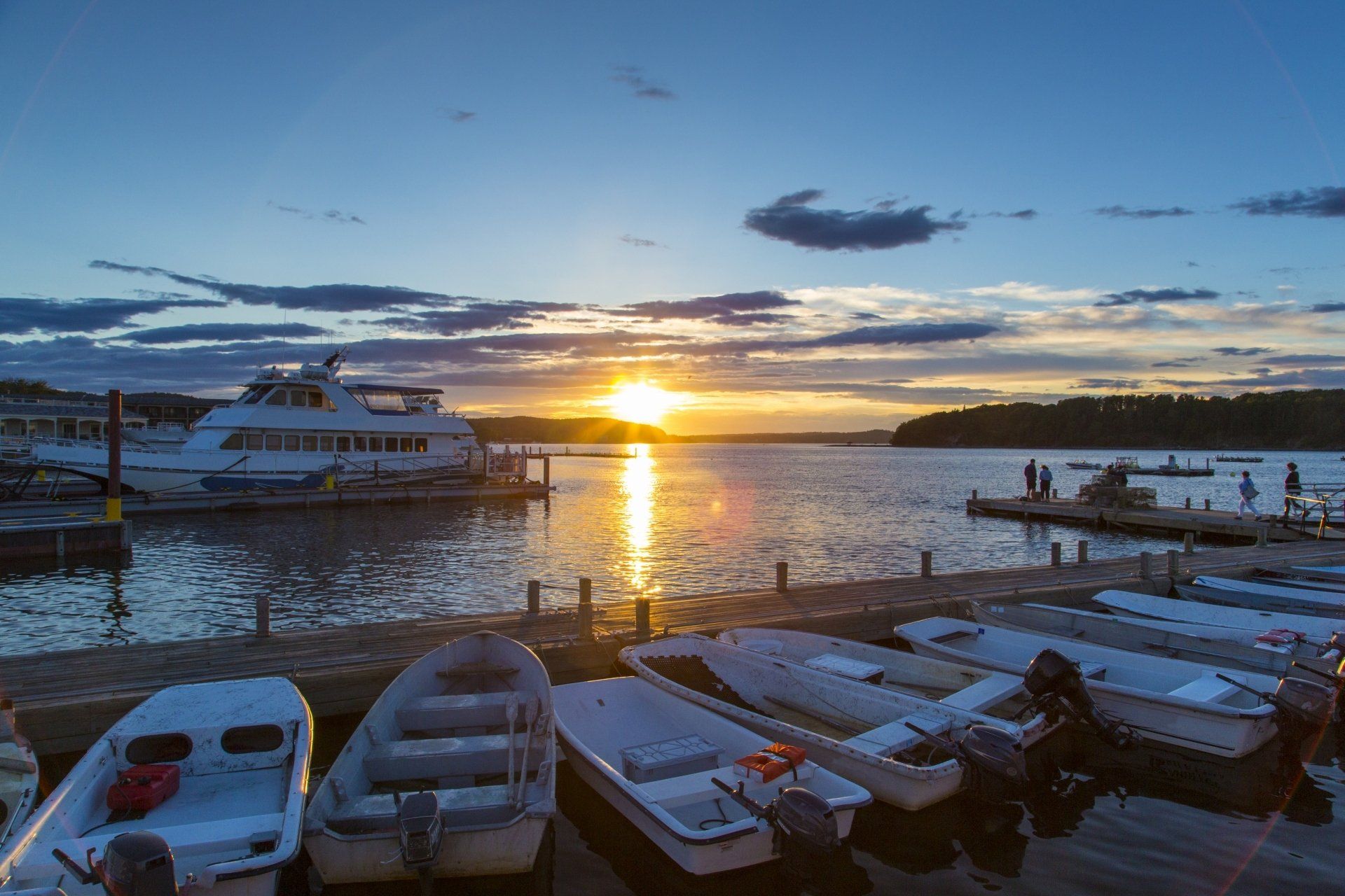 Zonsondergang boven de haven, kleine bootjes aangemeerd op de voorgrond, een grote boot aan de pier, een bewolkte hemel.