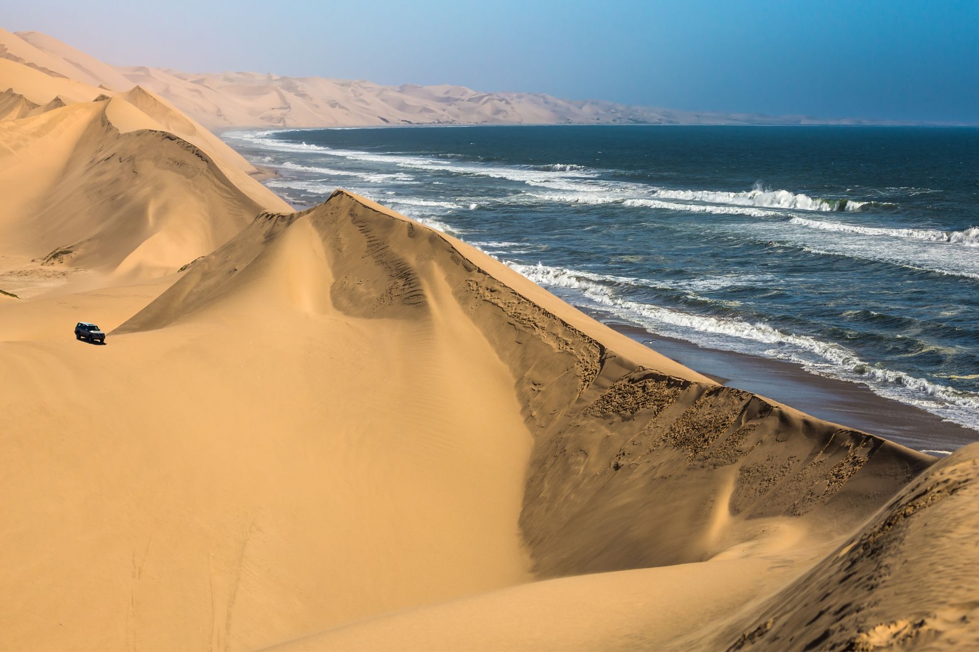 In Namibië lopen zandduinen door tot aan de oceaan; een voertuig staat op een duin.