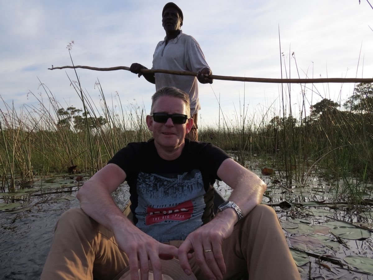 Man in een boot, kijkend naar de camera. Inheemse man staat erachter met een stok in zijn hand. Moerasgebied, zonsondergang.