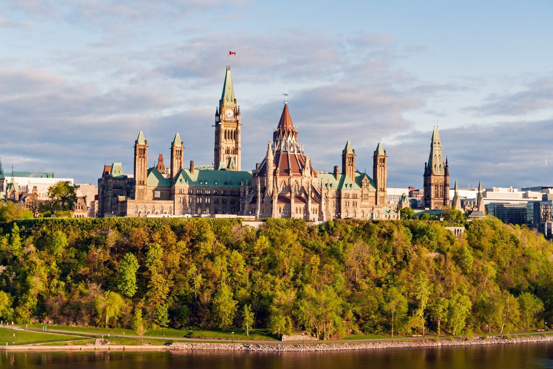 Het parlementsgebouw in Ottawa, Canada, staat op een met bomen begroeide heuvel met een rivier op de voorgrond.