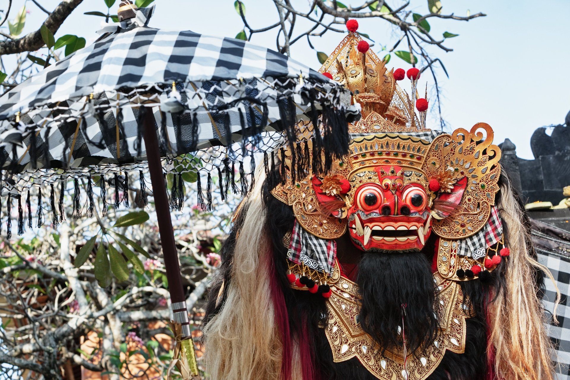 Balinese Barong-masker met gouden details en een geruite paraplu op de achtergrond.