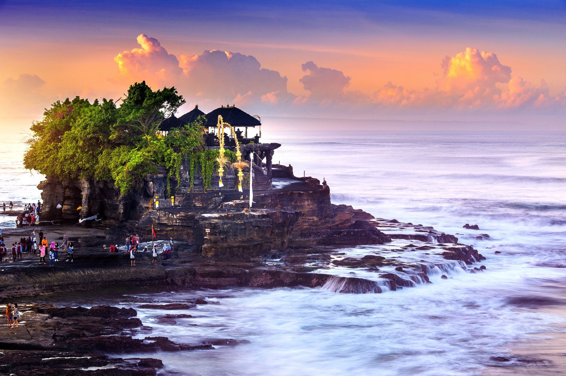 Tempel op een rotsachtig eiland, Bali, Indonesië, bij zonsondergang; golven breken op de kust, mensen in de buurt.