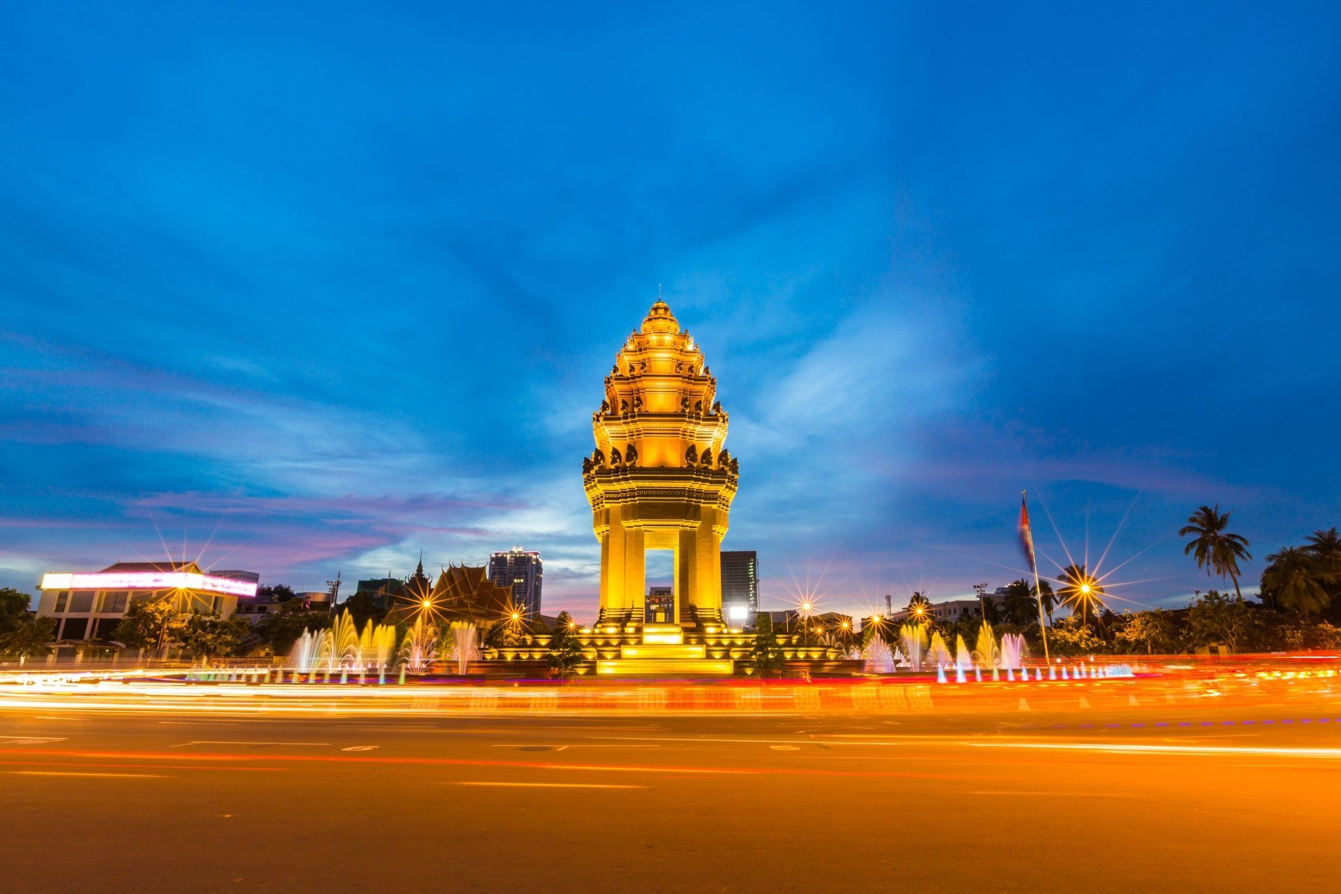 Monument 's nachts in Phnom Penh, Cambodja, met verlichte lampen en wazige autosporen, onder een schemerige hemel.