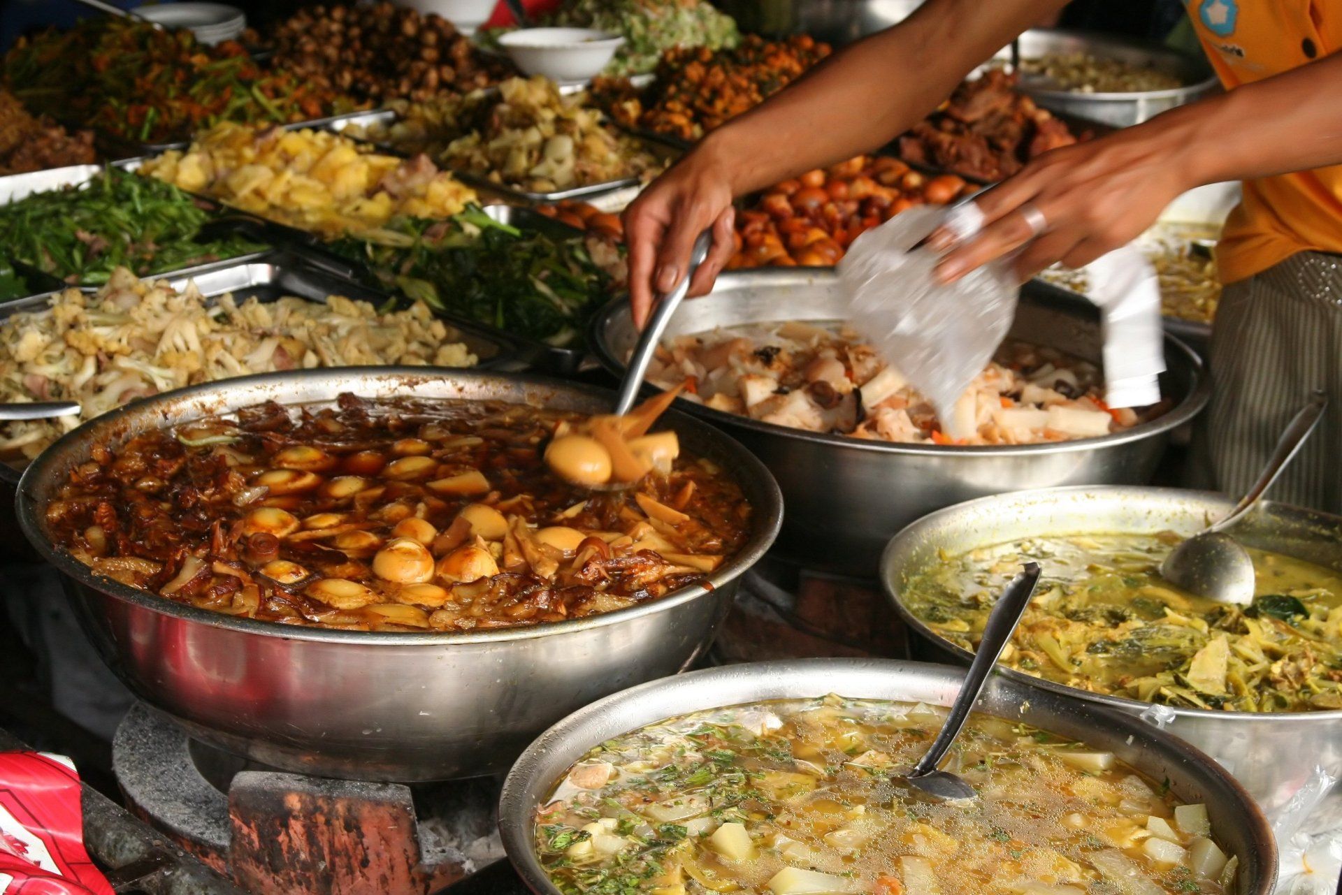 Iemand schept eten uit verschillende metalen potten bij een drukke eetkraam in de openlucht.