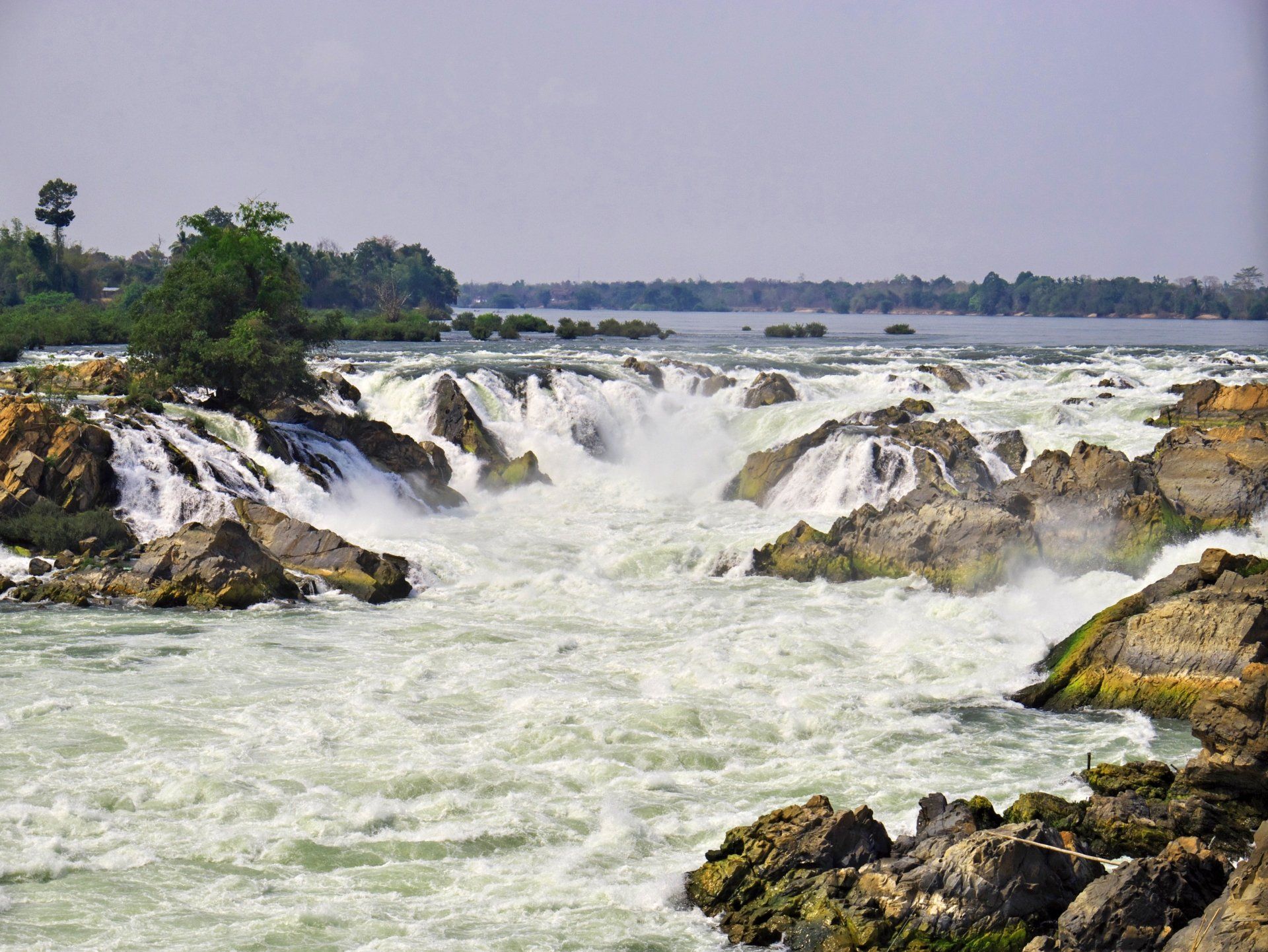 Kolkend wit water stort zich over rotsachtige watervallen in een brede rivier onder een wazige hemel.