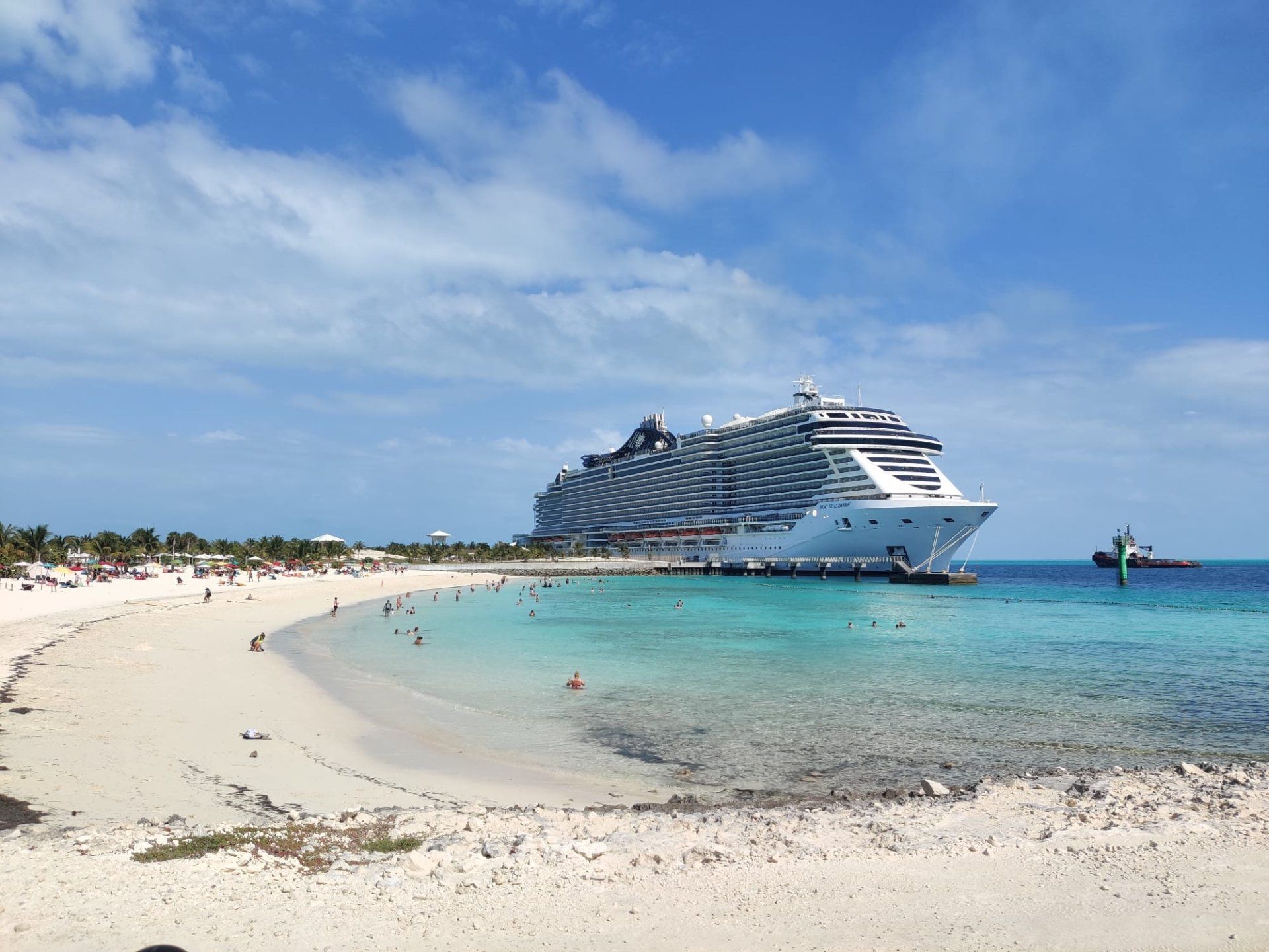 Strandtafereel met een groot cruiseschip aangemeerd; turkoois water, blauwe lucht, wit zand.
