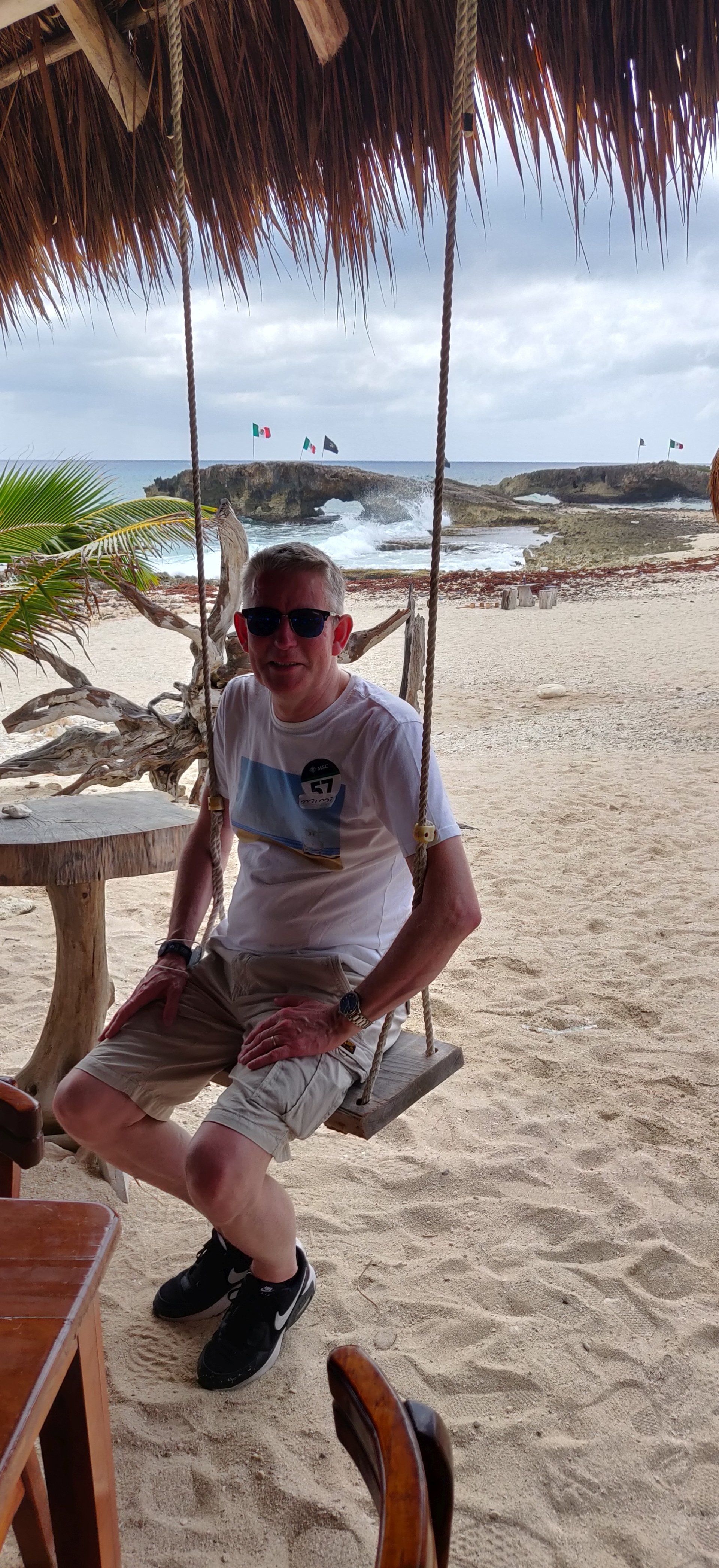 Een man schommelt op een zandstrand. Hij draagt een zonnebril en zit onder een rieten dak, met de oceaan op de achtergrond.