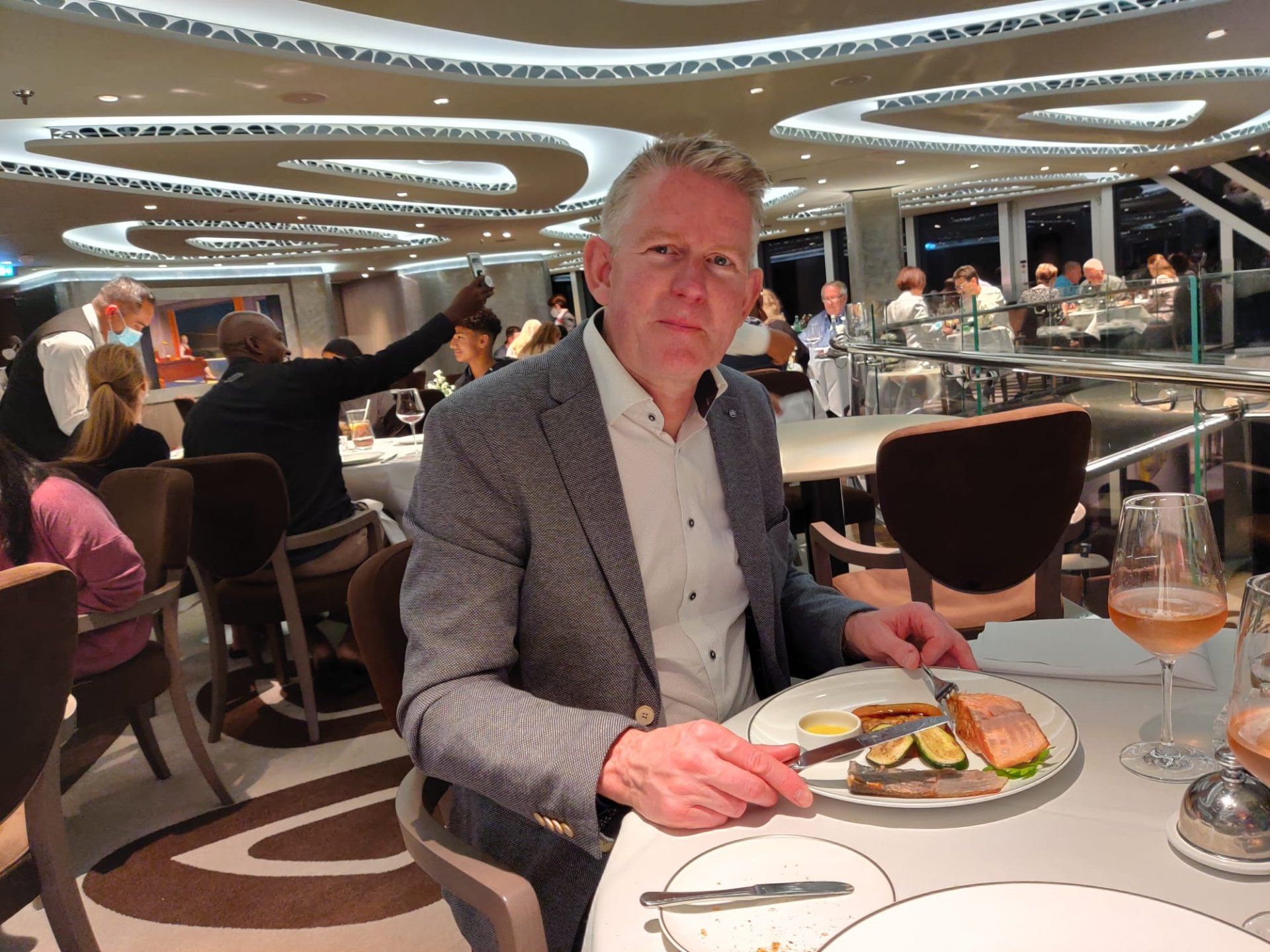 Een man eet in een restaurant. Hij draagt een blazer. Het eten ligt op een bord. Interieur met mensen op de achtergrond.