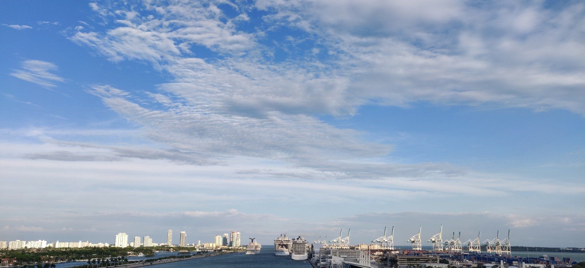 Een blauwe hemel met wolken boven een haven met schepen en in de verte de skyline van een stad.