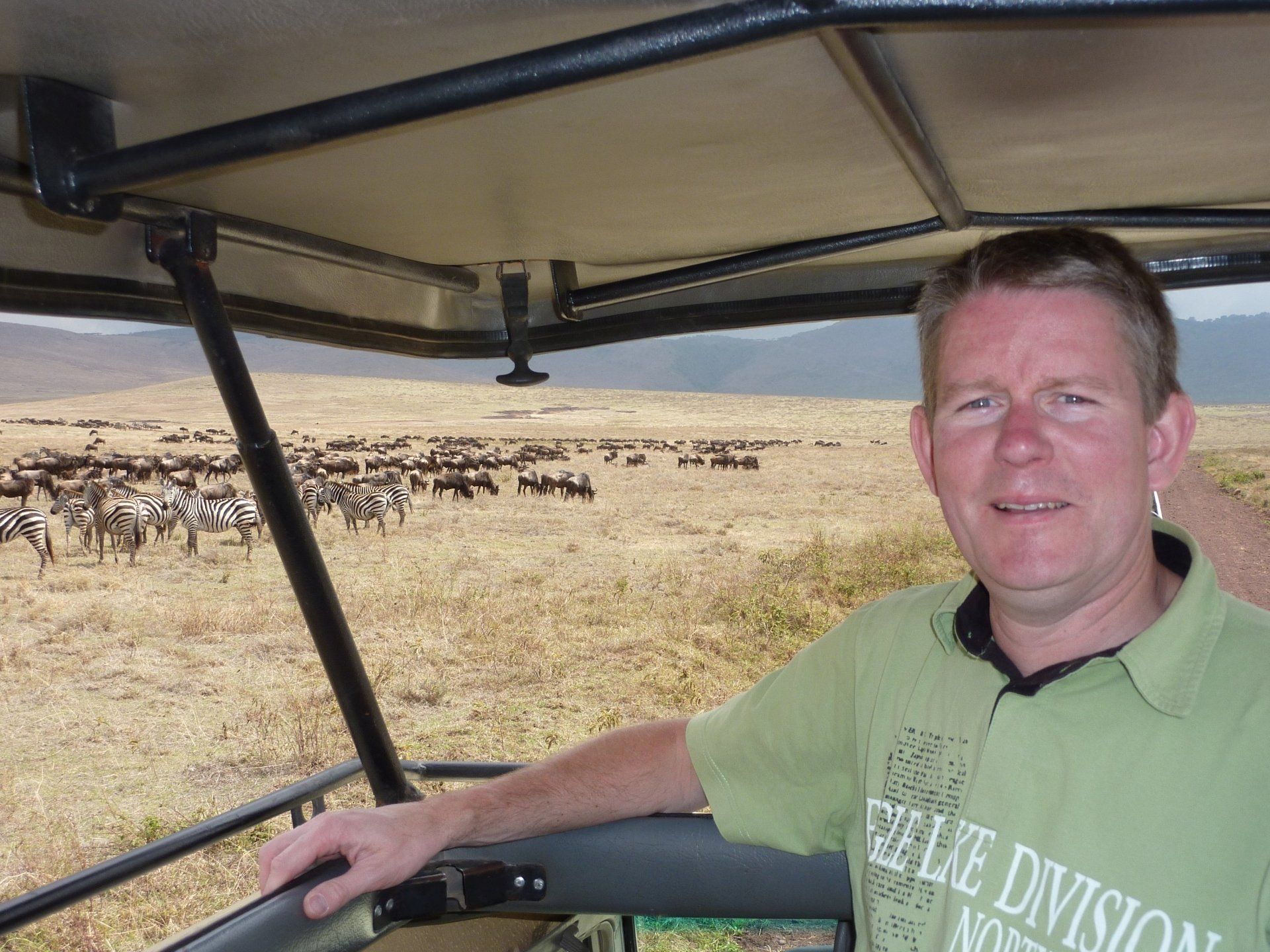 Een man in een safarivoertuig lacht naar de camera; op de achtergrond is een kudde zebra's en gnoes te zien.