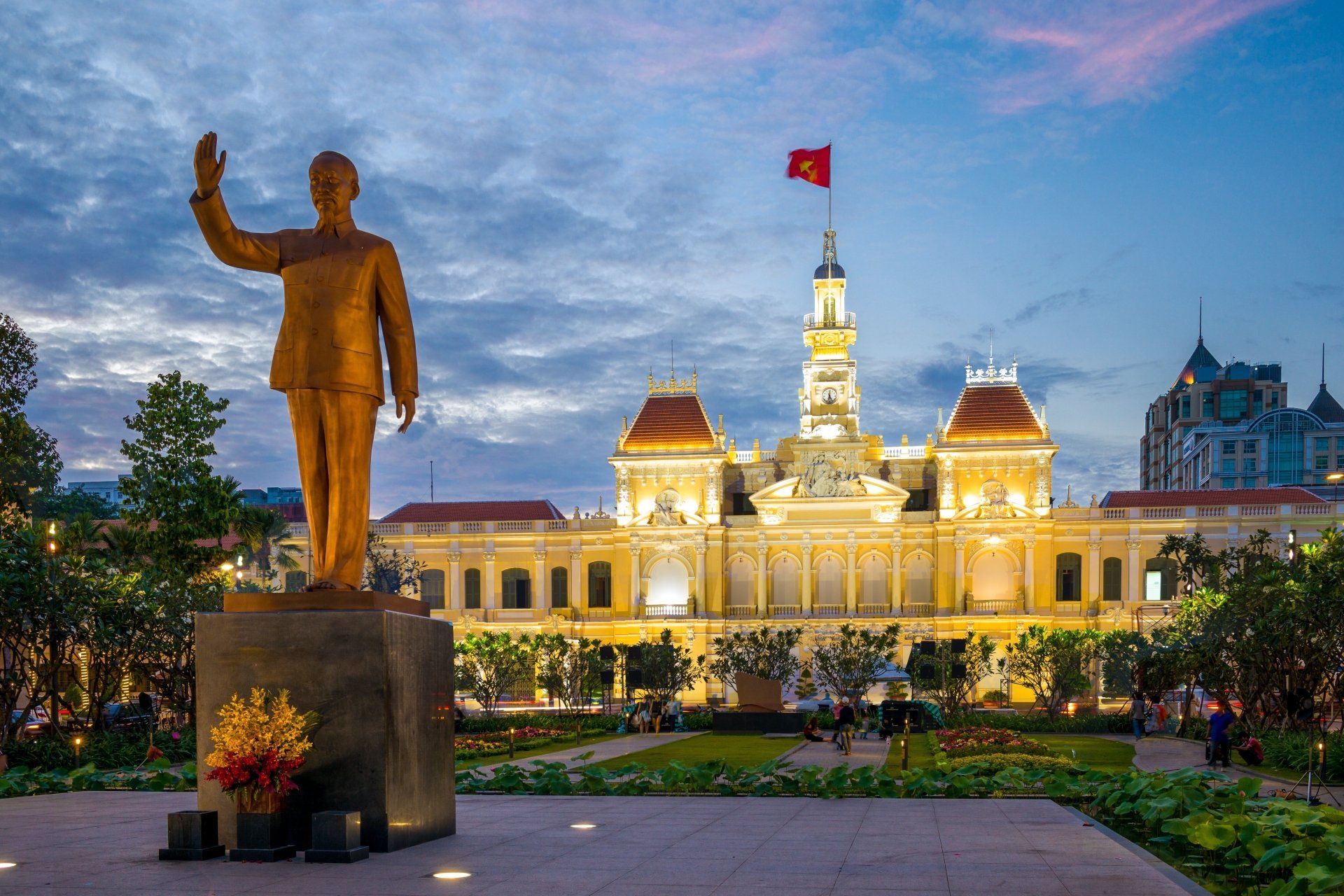 Standbeeld van Ho Chi Minh die zwaait voor het stadhuis van Ho Chi Minh-stad, verlicht in de schemering, met de Vietnamese vlag wapperend.