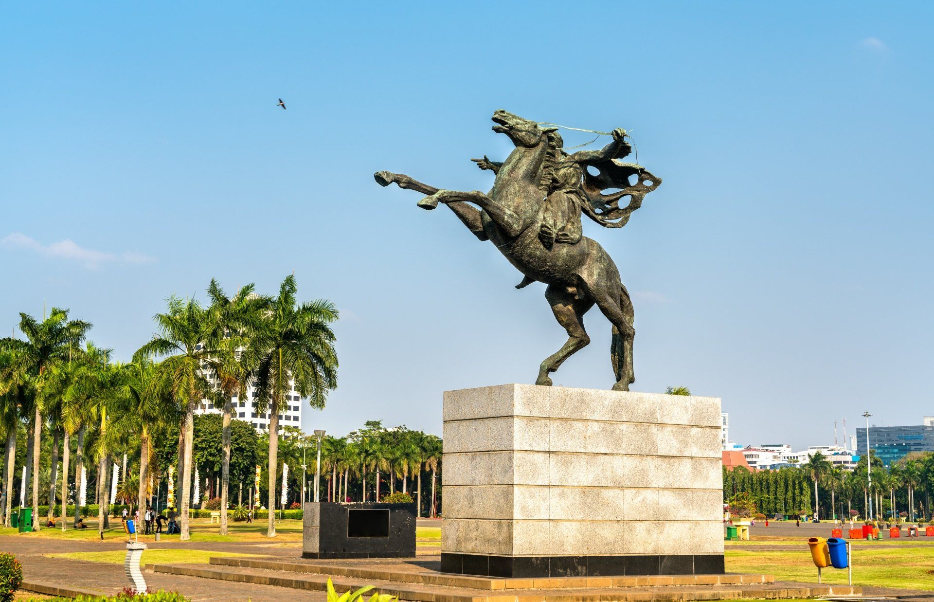 Bronzen ruiterbeeld op een stenen blok in een park, steigerend paard, palmbomen, blauwe lucht.