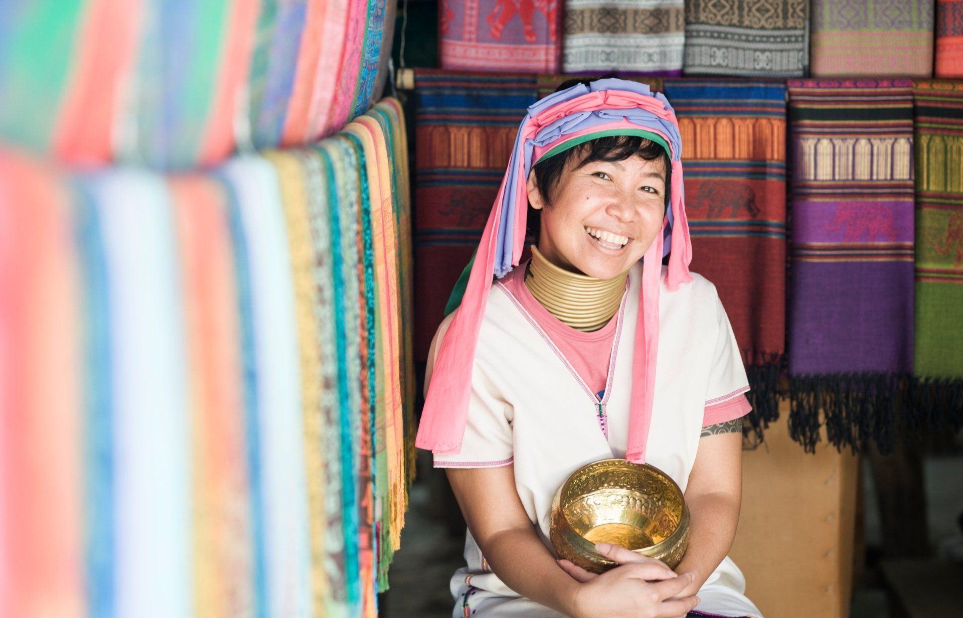 Een vrouw met traditionele halsringen en hoofdtooi glimlacht, terwijl ze een gouden kom vasthoudt, omringd door kleurrijke textielstoffen.