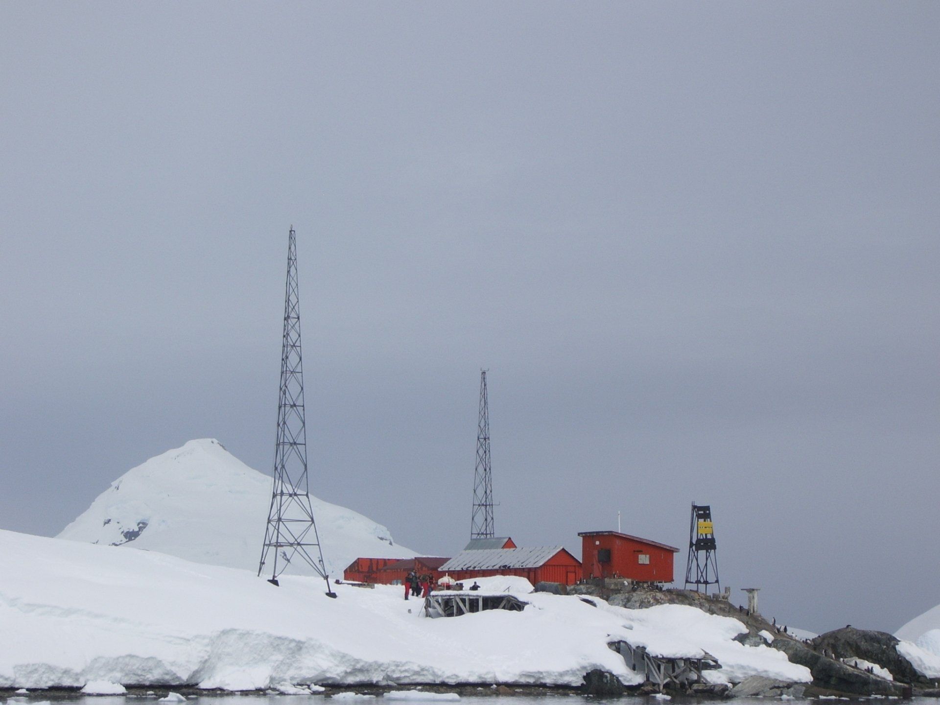 Rode gebouwen en radiotorens in een besneeuwd, bergachtig Antarctisch landschap onder een bewolkte hemel.