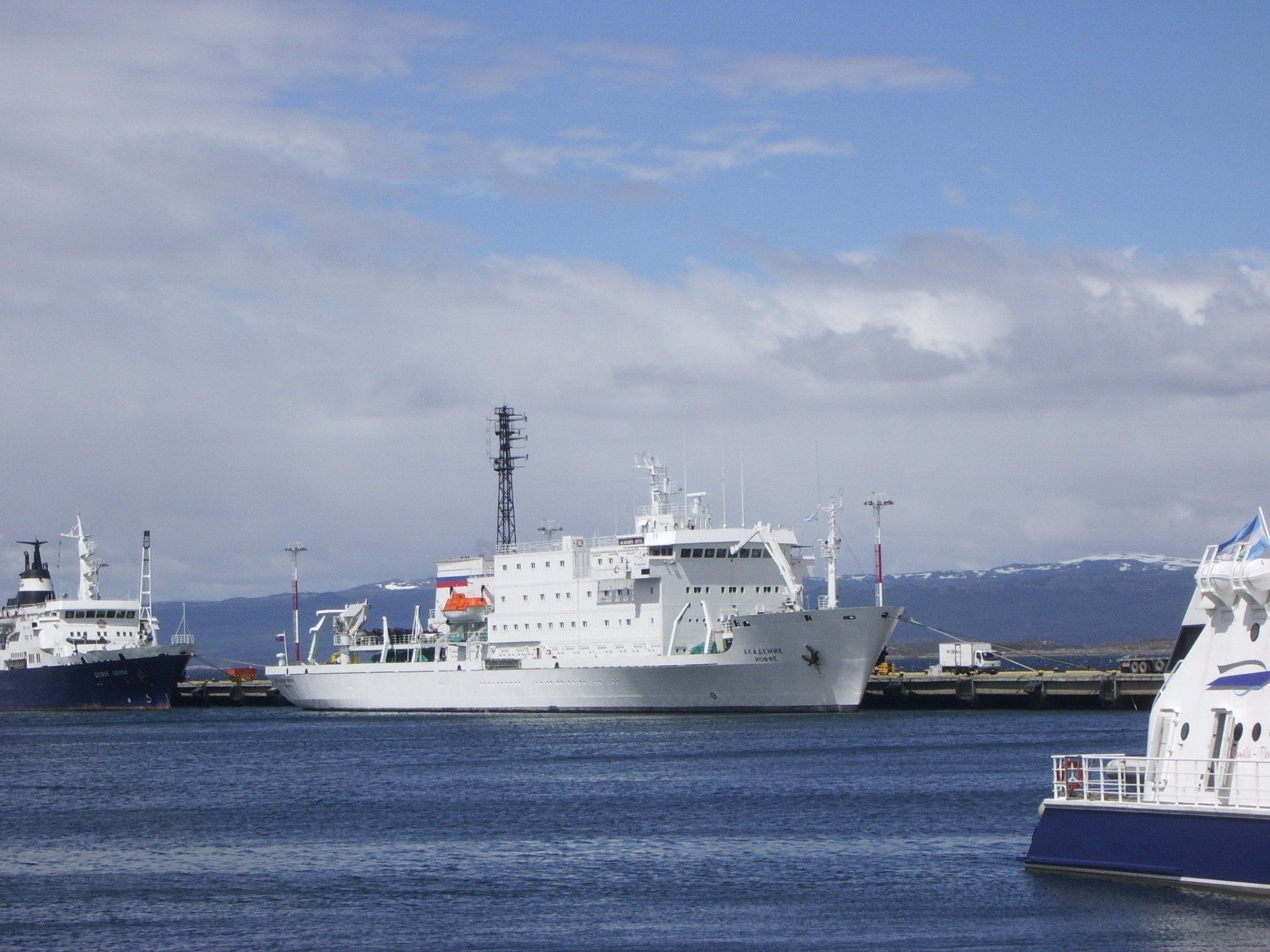 Wit onderzoeksschip aangemeerd in de haven, blauw water, bergen op de achtergrond.
