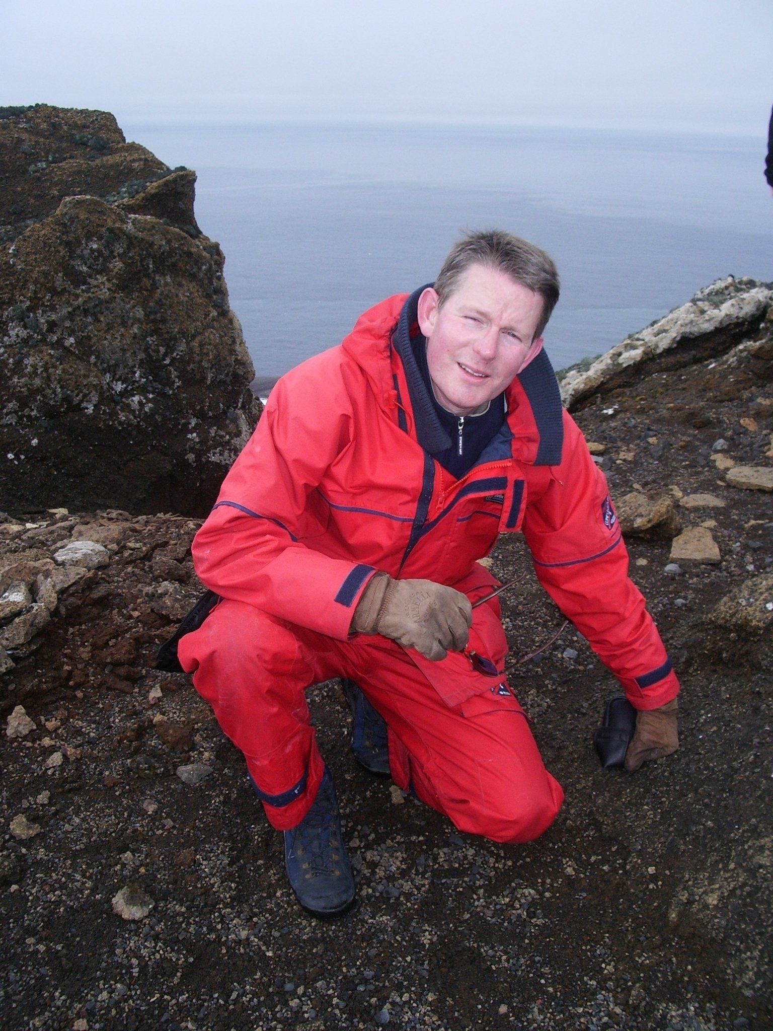 Een man in een rood overall hurkt op donker, rotsachtig terrein nabij een waterpartij, draagt handschoenen en kijkt naar beneden.