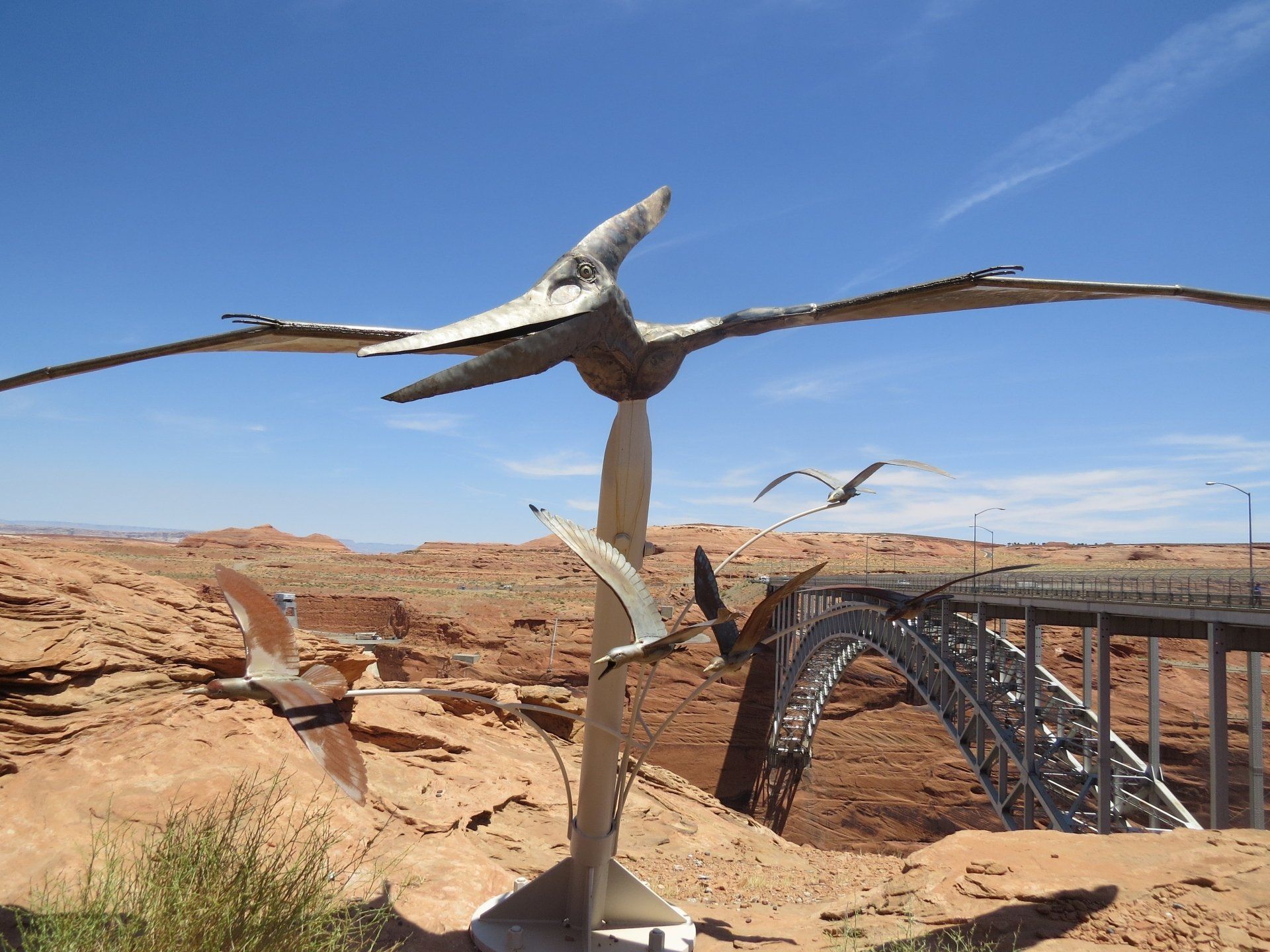 Metalen pterodactylussculpturen met een brug en een canyon op de achtergrond. Helderblauwe lucht.