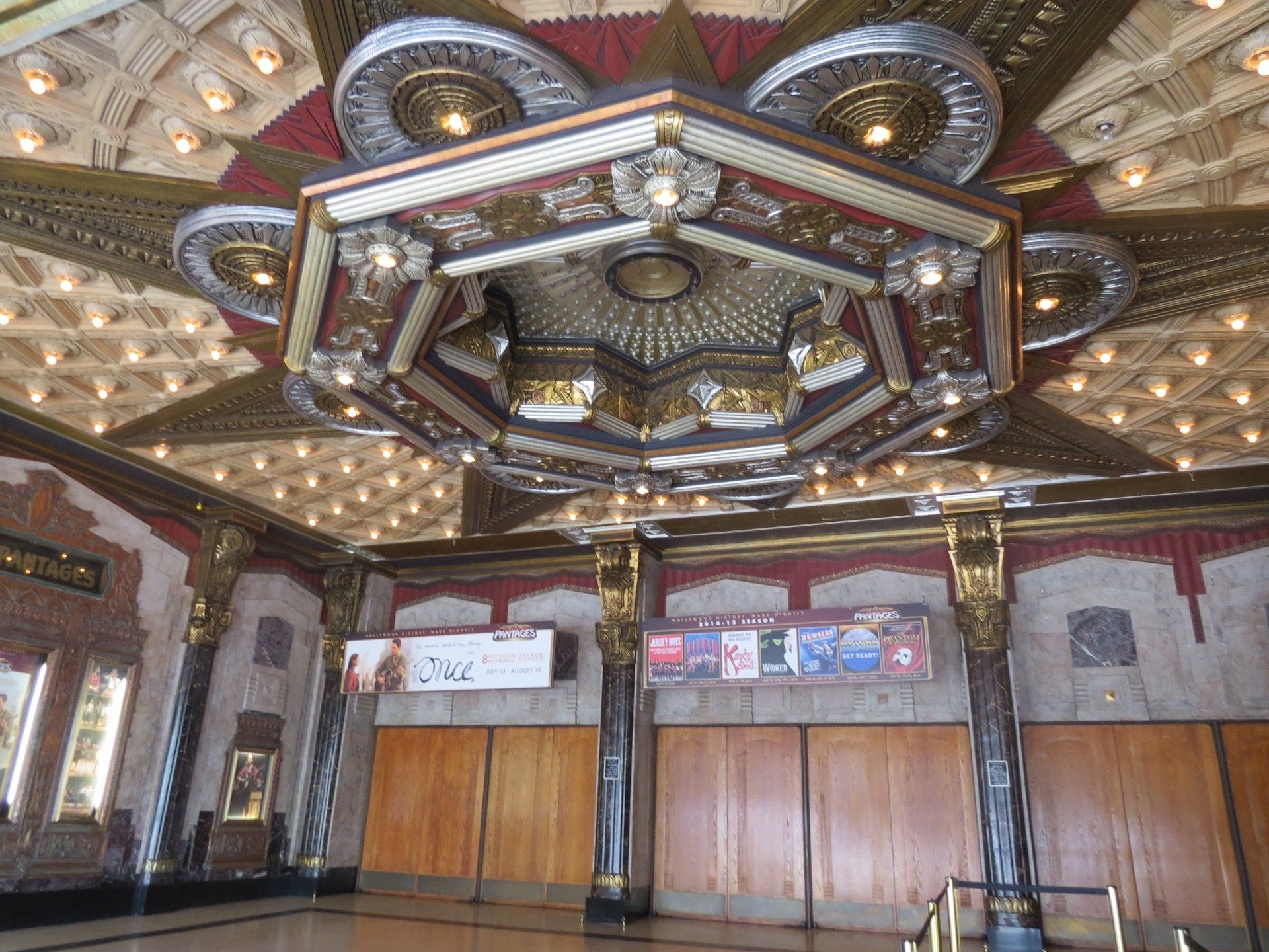 Een rijkversierde theaterlobby met een fraai plafond, houten deuren en posters.