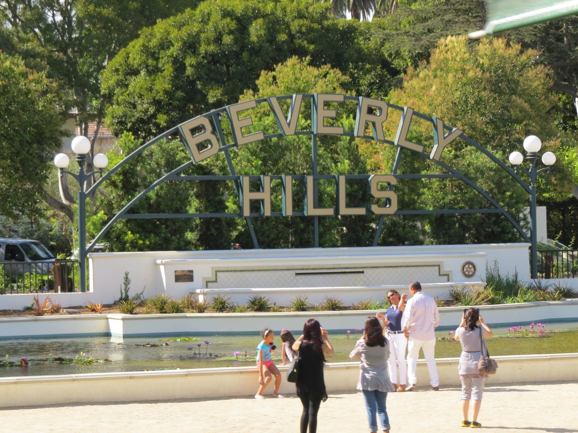 Een boog met het opschrift Beverly Hills hangt boven een reflecterend waterbassin, waar verschillende mensen foto's maken.