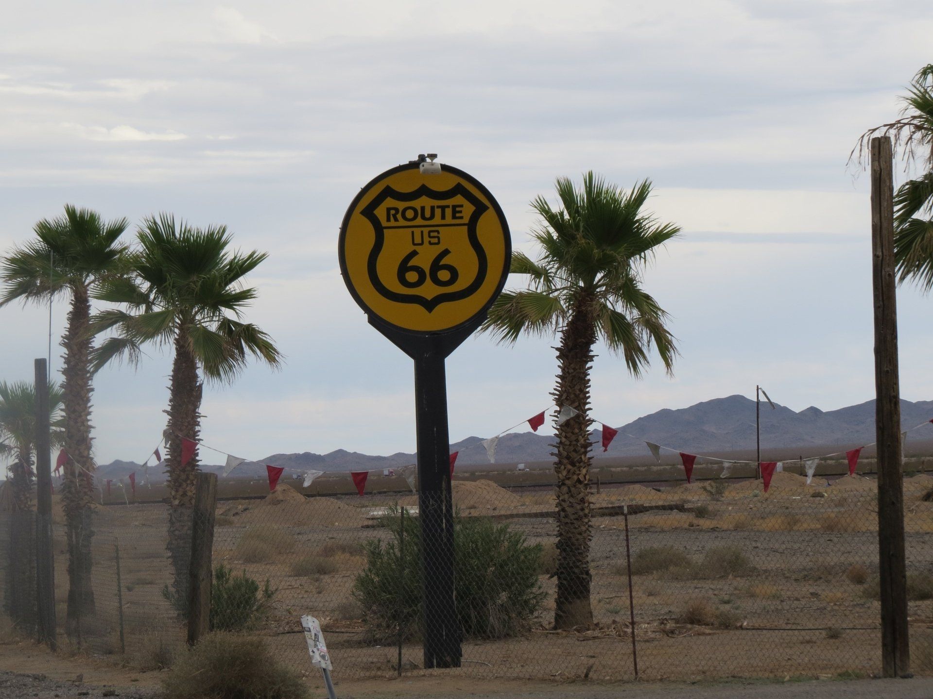Een Route 66-bord op een zwarte paal, omringd door palmbomen, een woestijnlandschap en een gaashek.