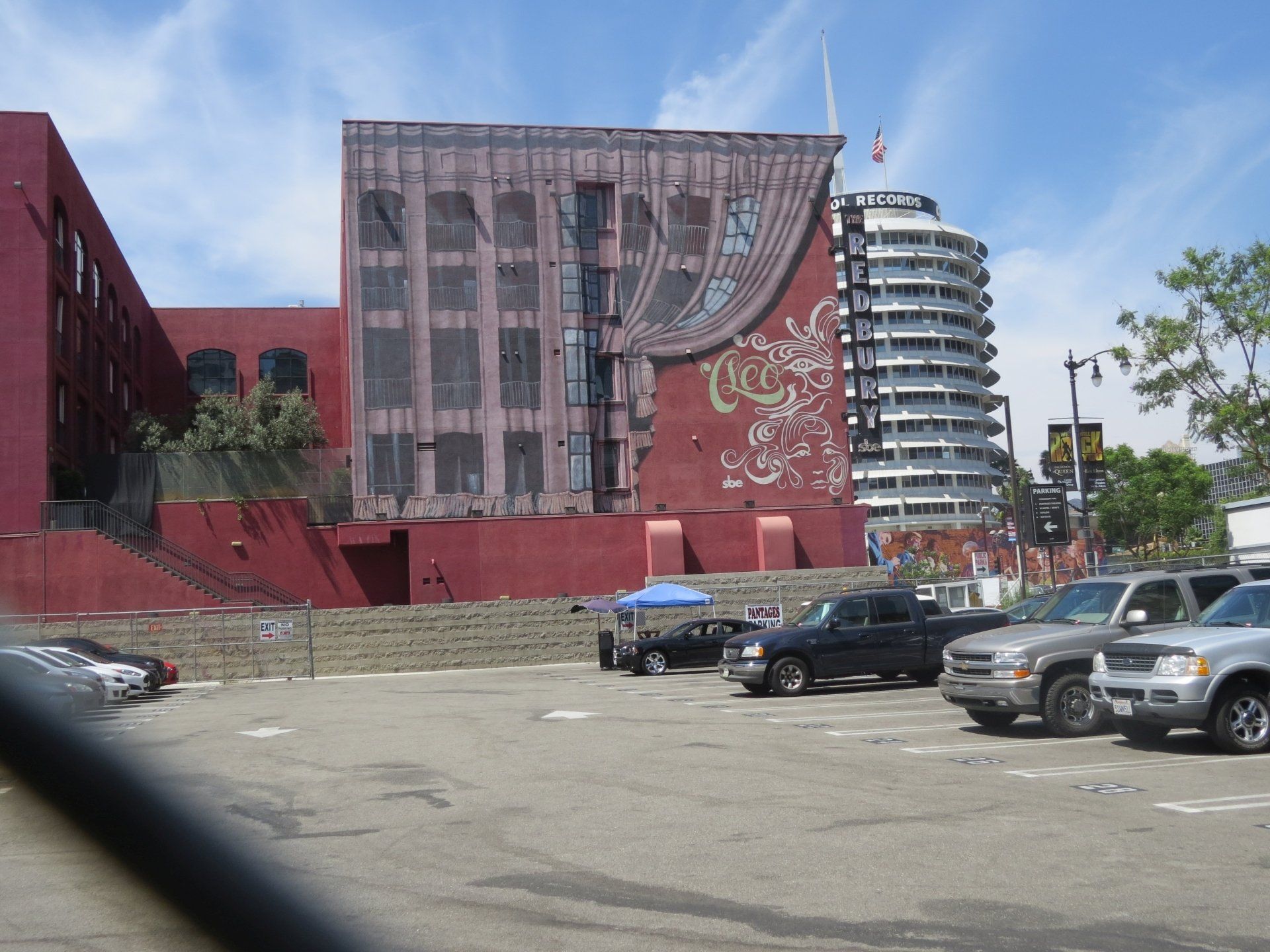 Een rood bakstenen gebouw met een muurschildering op een parkeerplaats in Hollywood, Californië, met op de achtergrond een hoog, rond gebouw.
