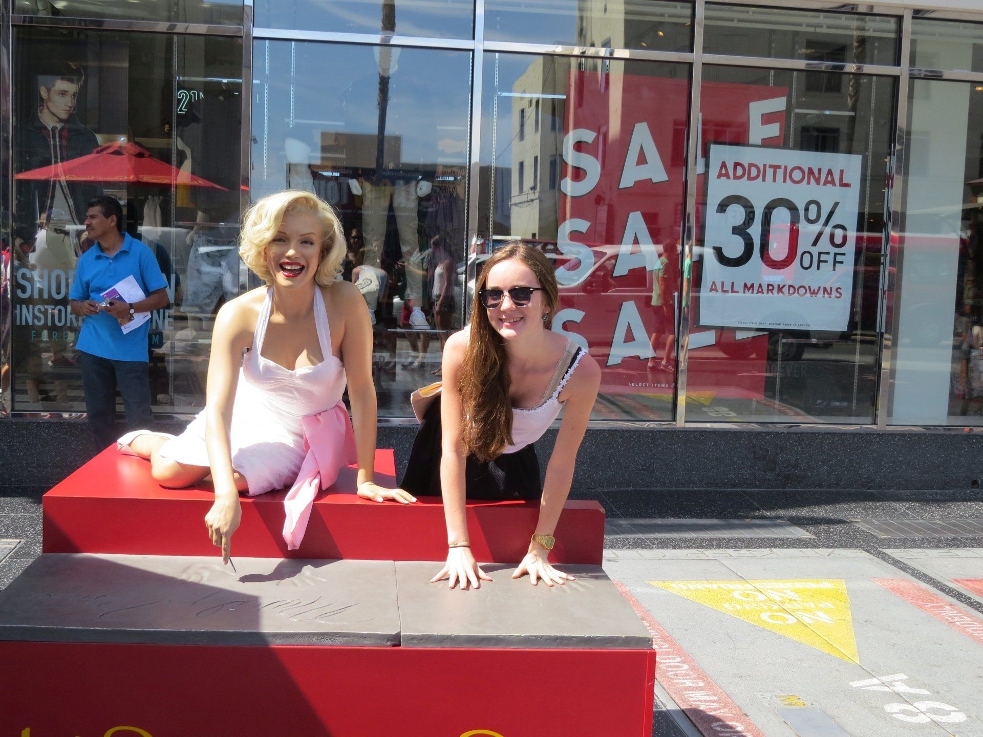 Twee vrouwen poseren op een rood podium op de Hollywood Walk of Fame. De ene draagt een Marilyn Monroe-kostuum, de andere een zonnebril. Achter hen bevindt zich een winkel.