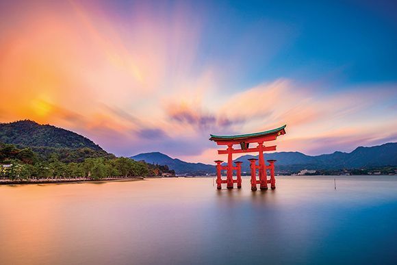 Rode torii-poort in het water bij zonsondergang, met bergen en een kleurrijke hemel op de achtergrond.