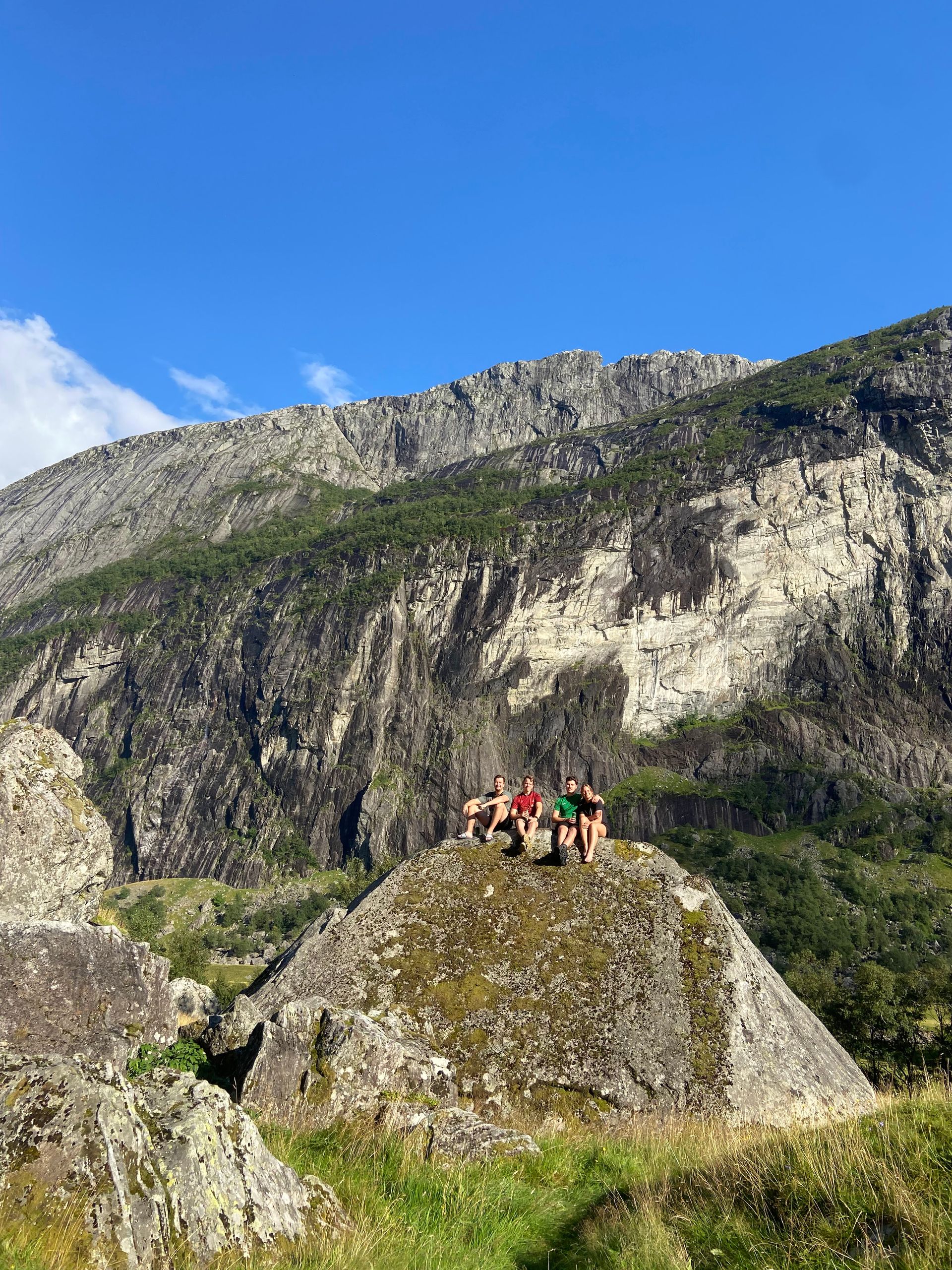 Een groep mensen poseert op een grote rots met een berg op de achtergrond. Groene vegetatie en een blauwe lucht.