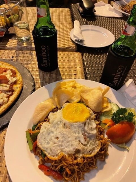Een bord gebakken noedels met een gebakken ei, crackers en groenten, naast bier en pizza.