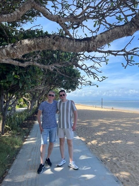 Twee mannen staan op een pad langs het strand. De ene draagt een gestreept shirt en een korte broek; de andere een gestreept shirt en een spijkerbroek.