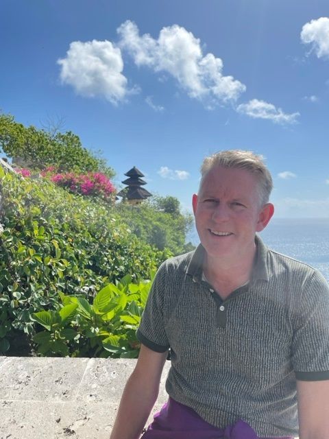 Een lachende man, gekleed in een groen shirt en paarse sarong, staat voor een tempel met uitzicht op de oceaan, Bali.