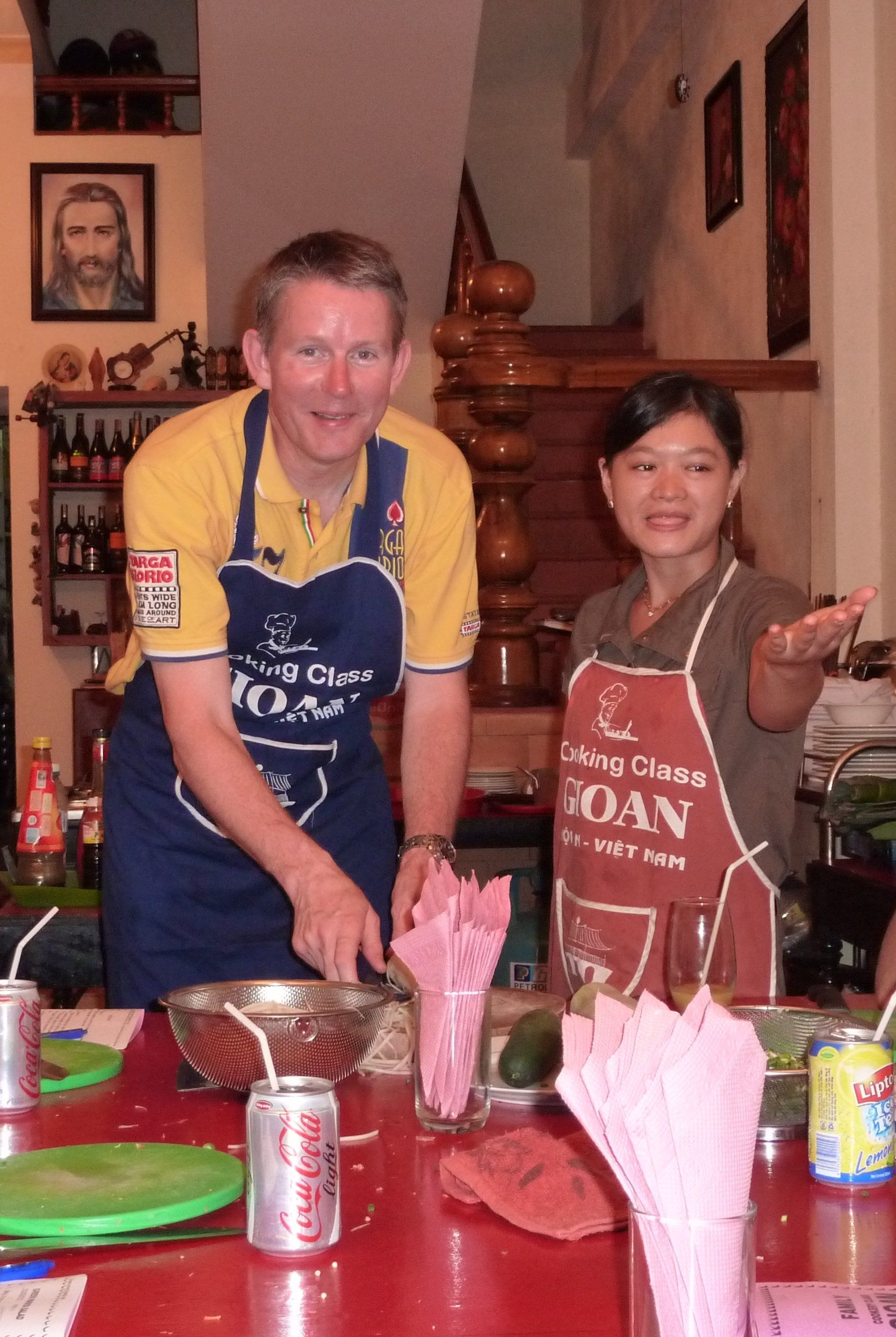 Een man en een vrouw in schorten bereiden eten in een keuken. Ze staan ​​bij een rode tafel en glimlachen.