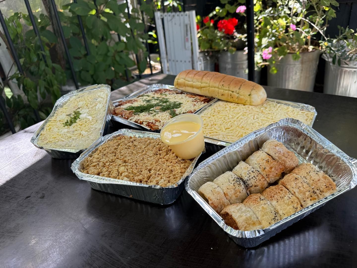 Prepared food trays on a dark table: lasagna, breadsticks, rolls, cheese, a side of sauce, and garlic bread, outdoors.