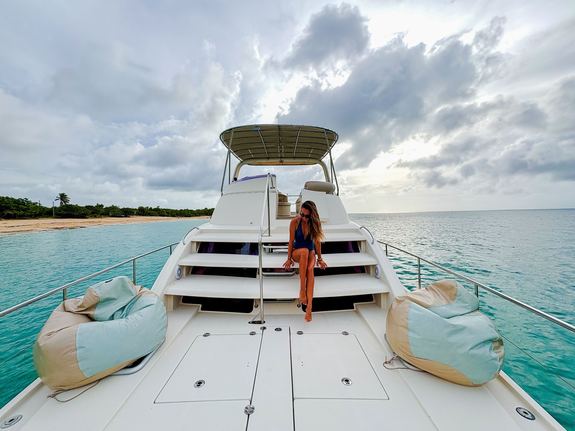 Woman sits on yacht steps, turquoise water, beach in the distance, cloudy sky.