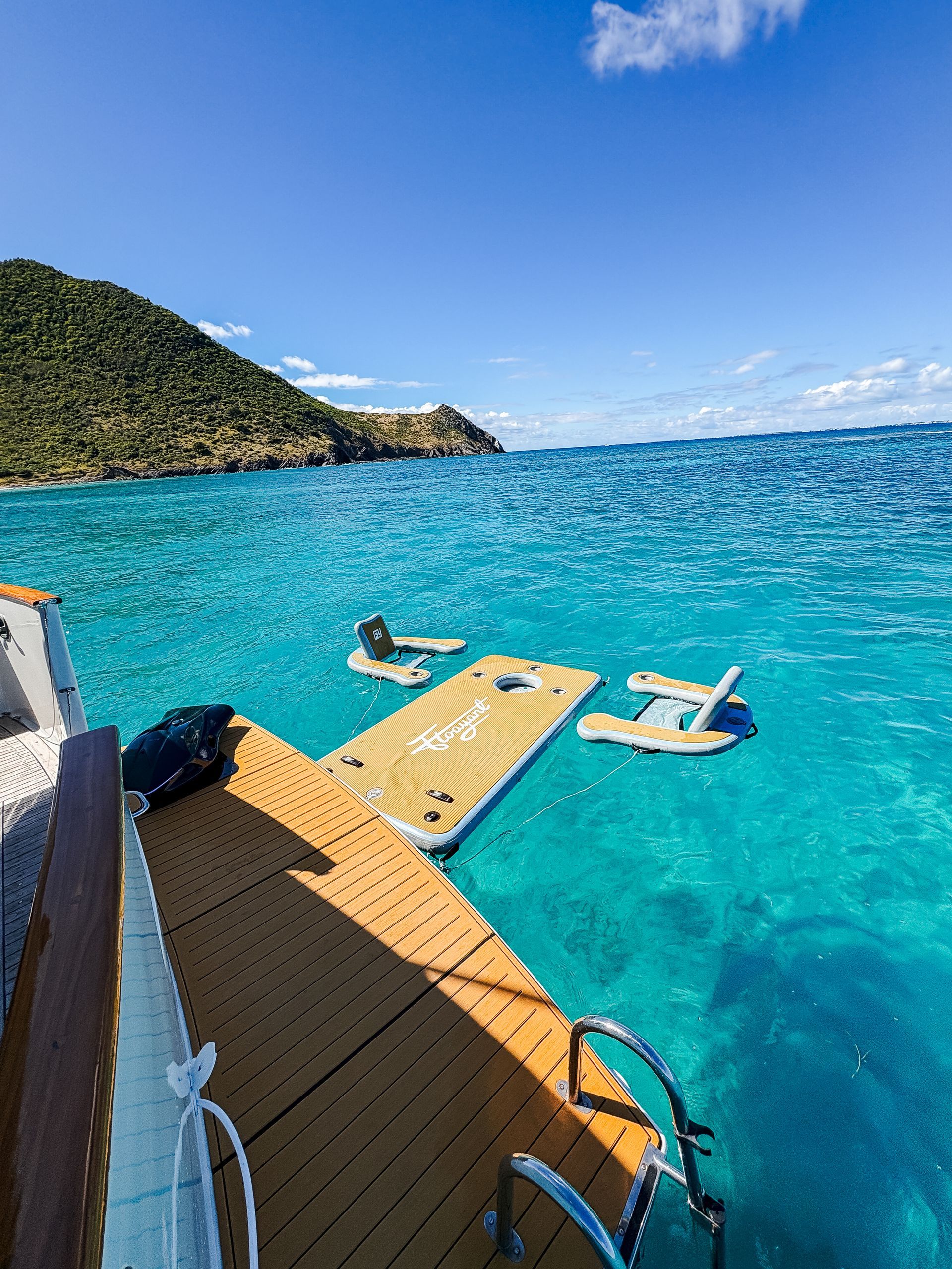 Wooden dock extending into turquoise water with floating platforms; hill in the background under a blue sky.