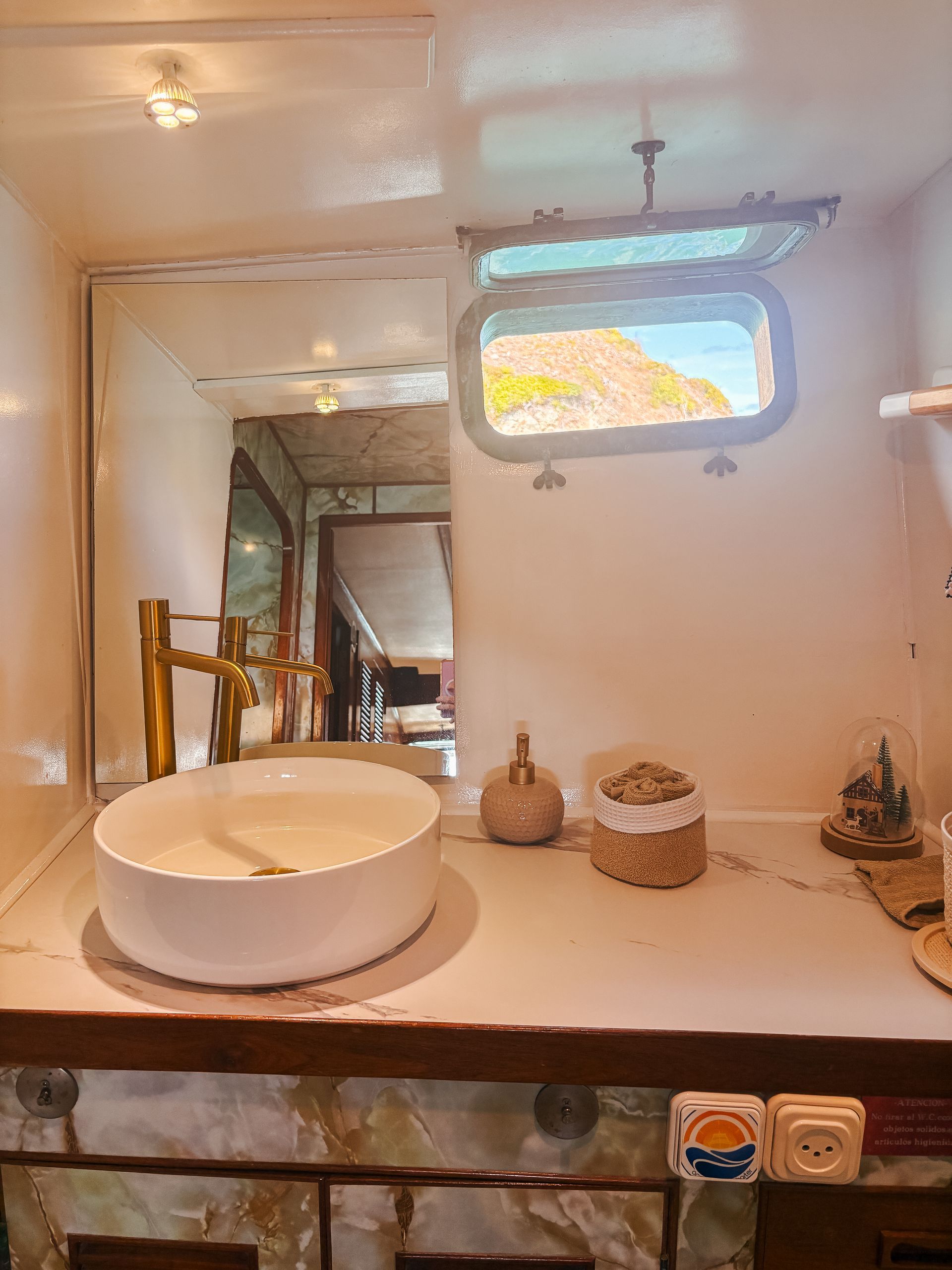 Bathroom with white sink, gold faucet, porthole window, large mirror, stone countertop.